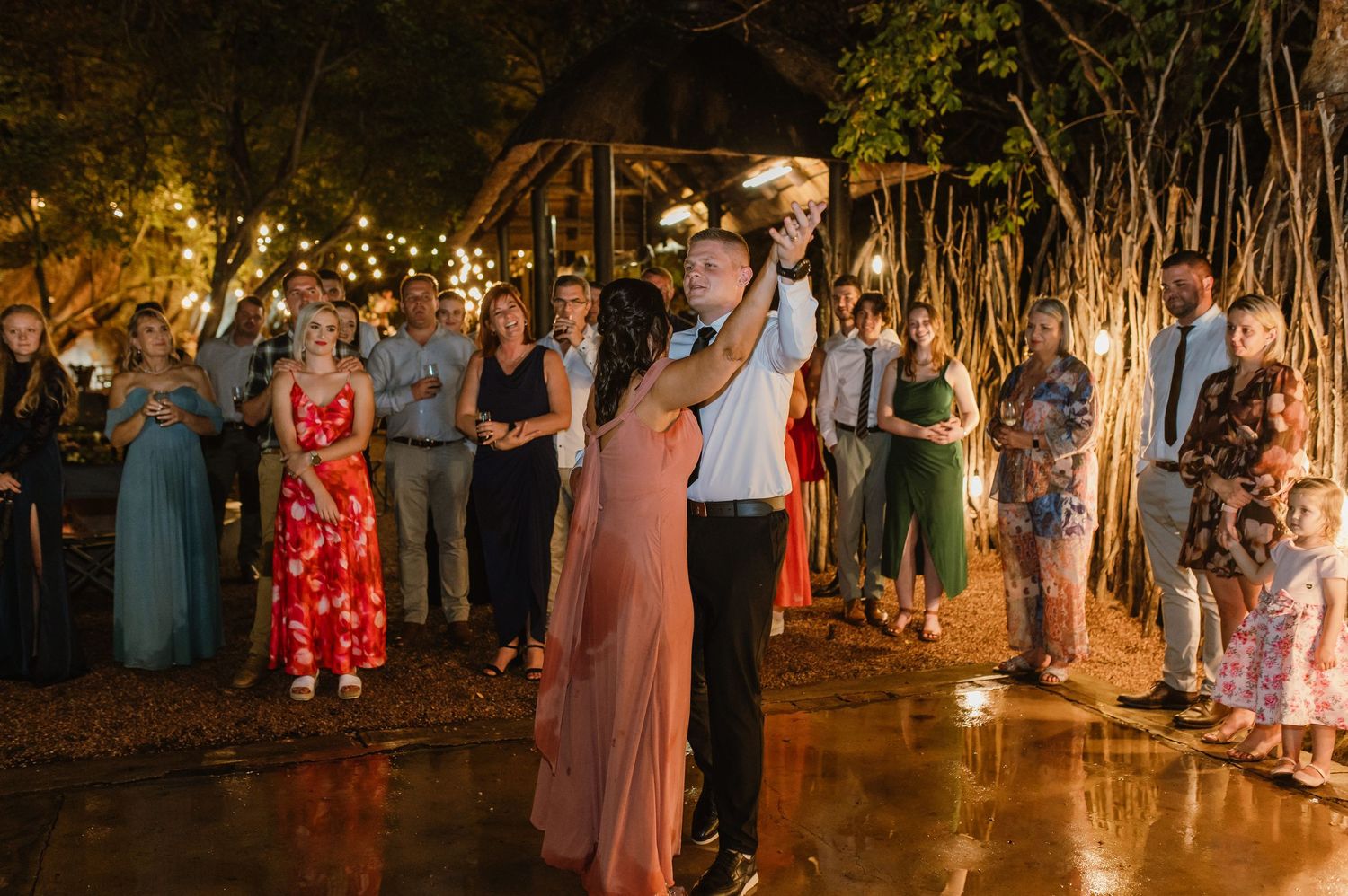 Couple dancing outdoors at night wedding reception surrounded by guests and sparkling string lights.