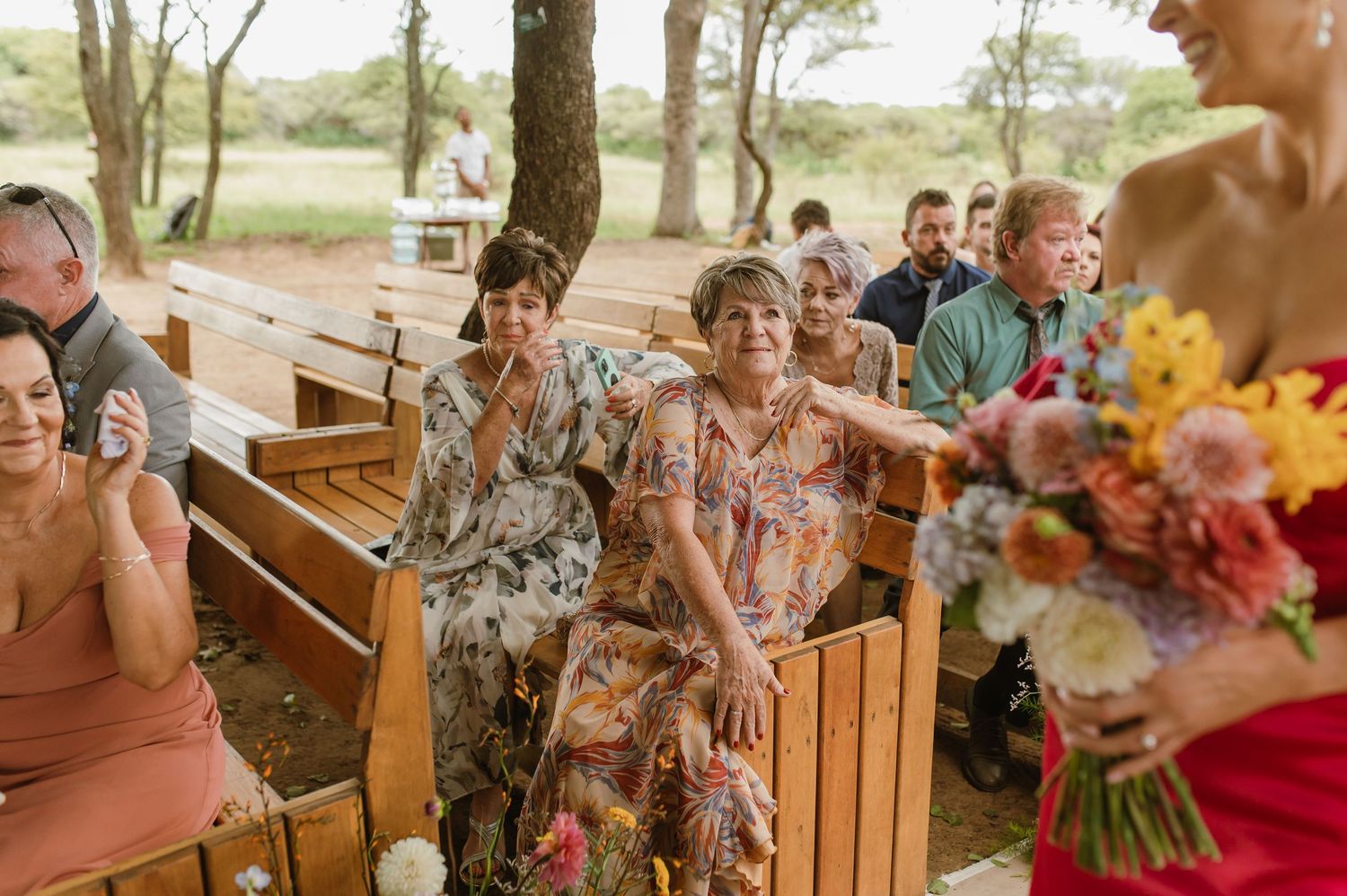 Emotional wedding guests seated on wooden benches watching bride walk down outdoor ceremony aisle.