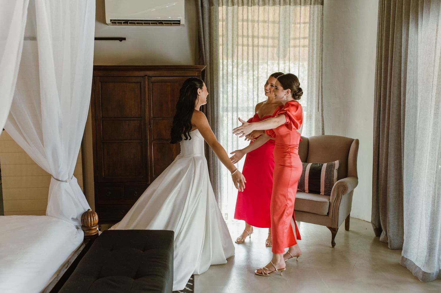 Bride in white gown embracing a woman in red dress in an elegant hotel room before wedding.