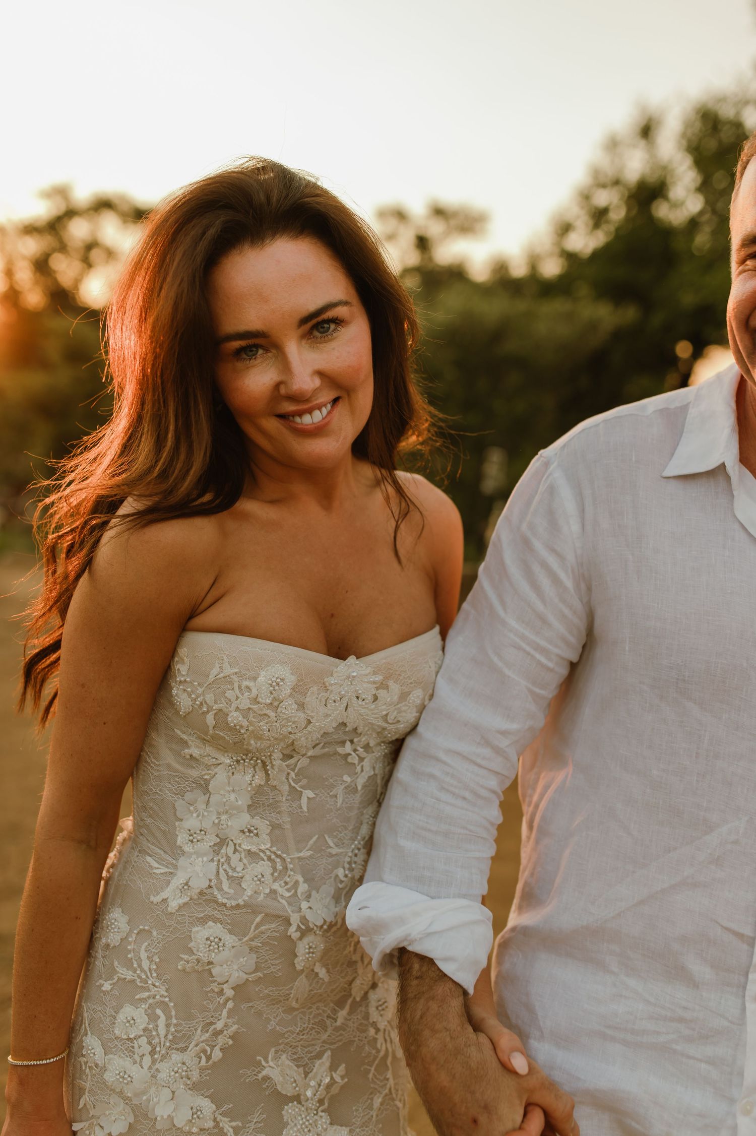 Radiant bride in strapless lace wedding gown smiles while holding hands with groom at sunset.