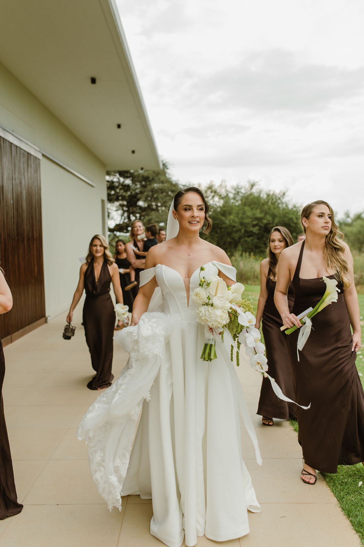 Wedding party walking down covered walkway with bride in white gown and bridesmaids in brown dresses.
