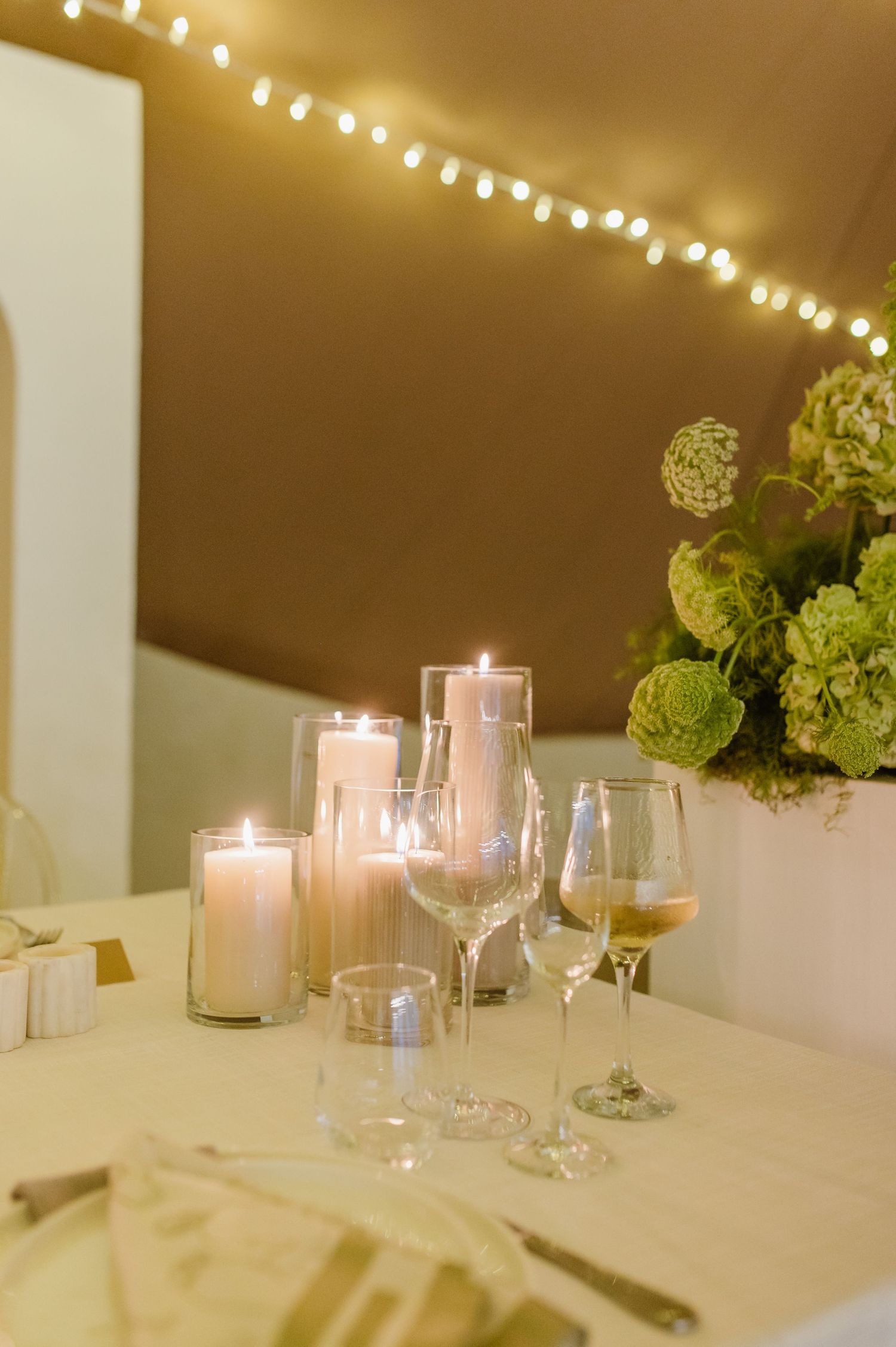 Elegant wedding reception table setting with white linens, glowing candles and green floral centerpieces.