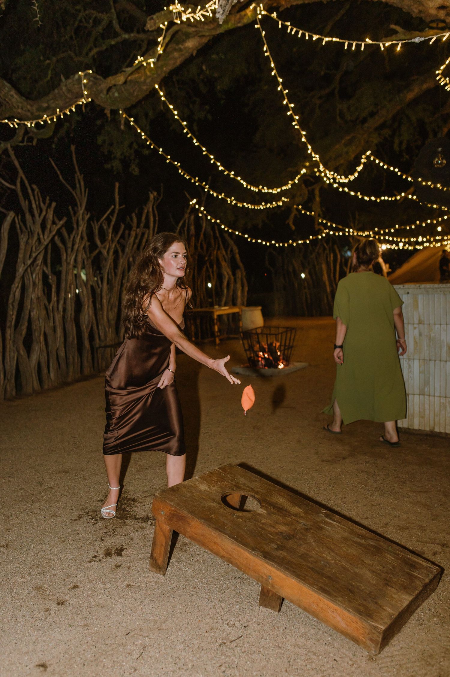 Guest playing cornhole game under string lights at nighttime outdoor wedding reception.