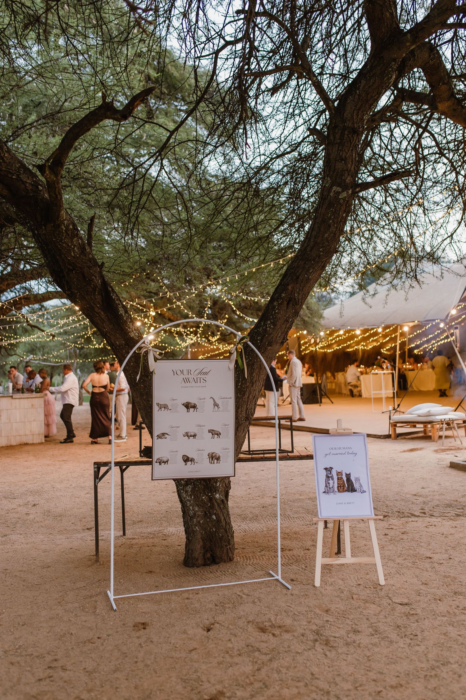 Wedding signage displayed on stands beneath a tree decorated with string lights at dusk.