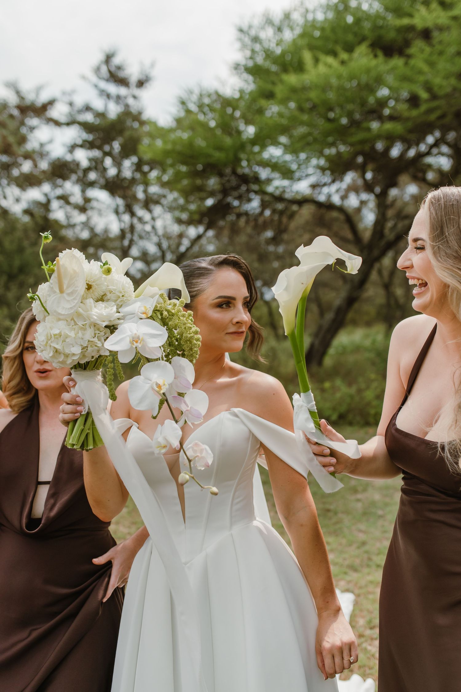 Bridal party holding white floral bouquets while laughing together outdoors.