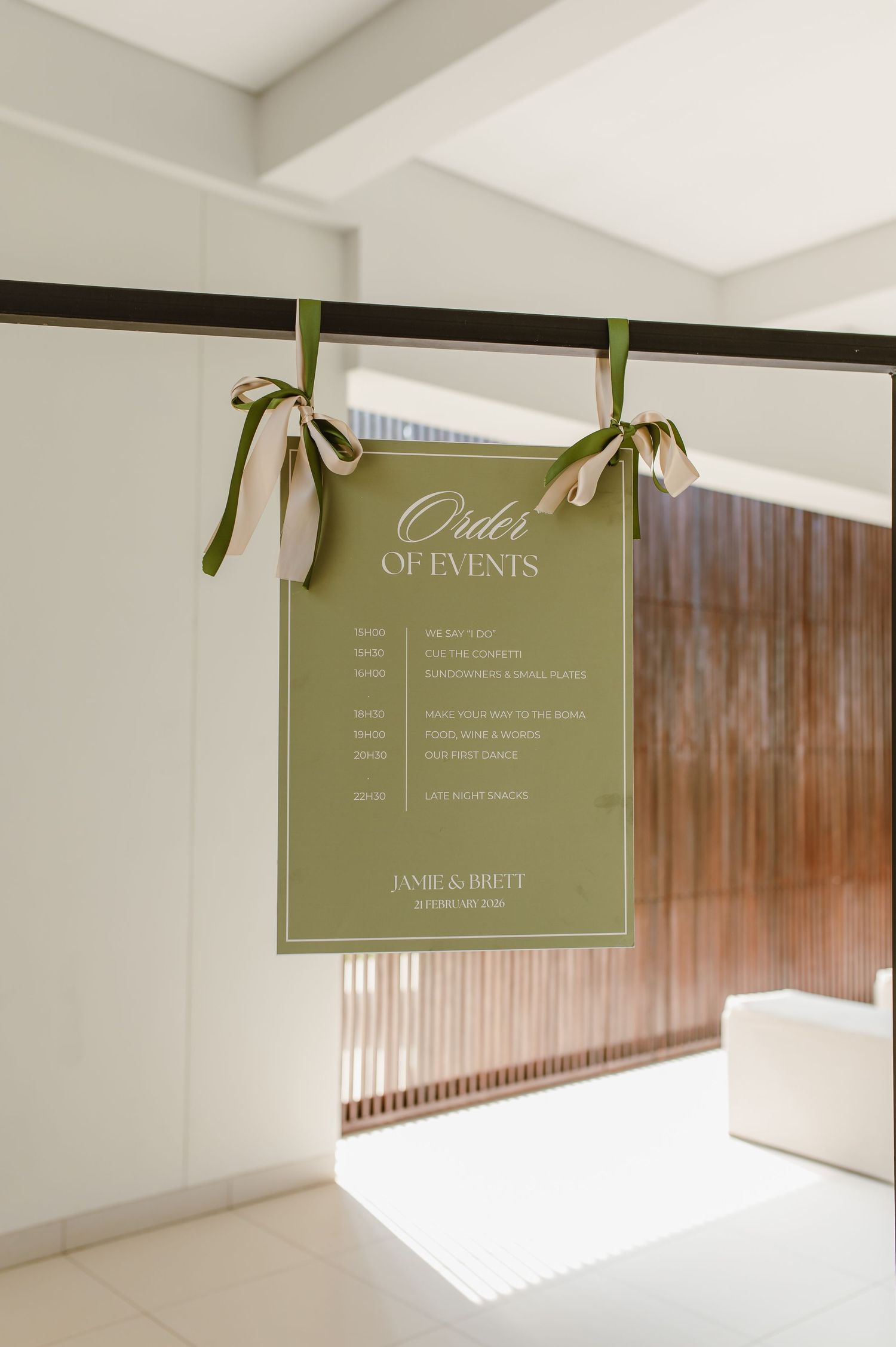 Elegant olive green wedding order of events sign hanging with ribbons against a white wall.