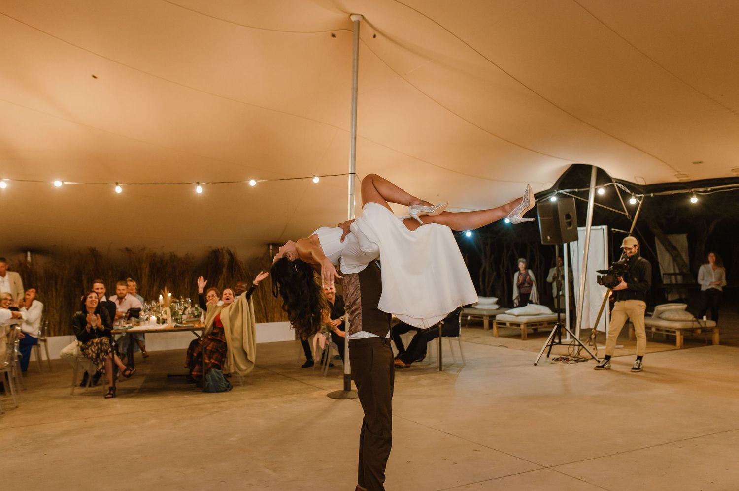 Wedding reception dance floor with guests celebrating under string lights in tent.
