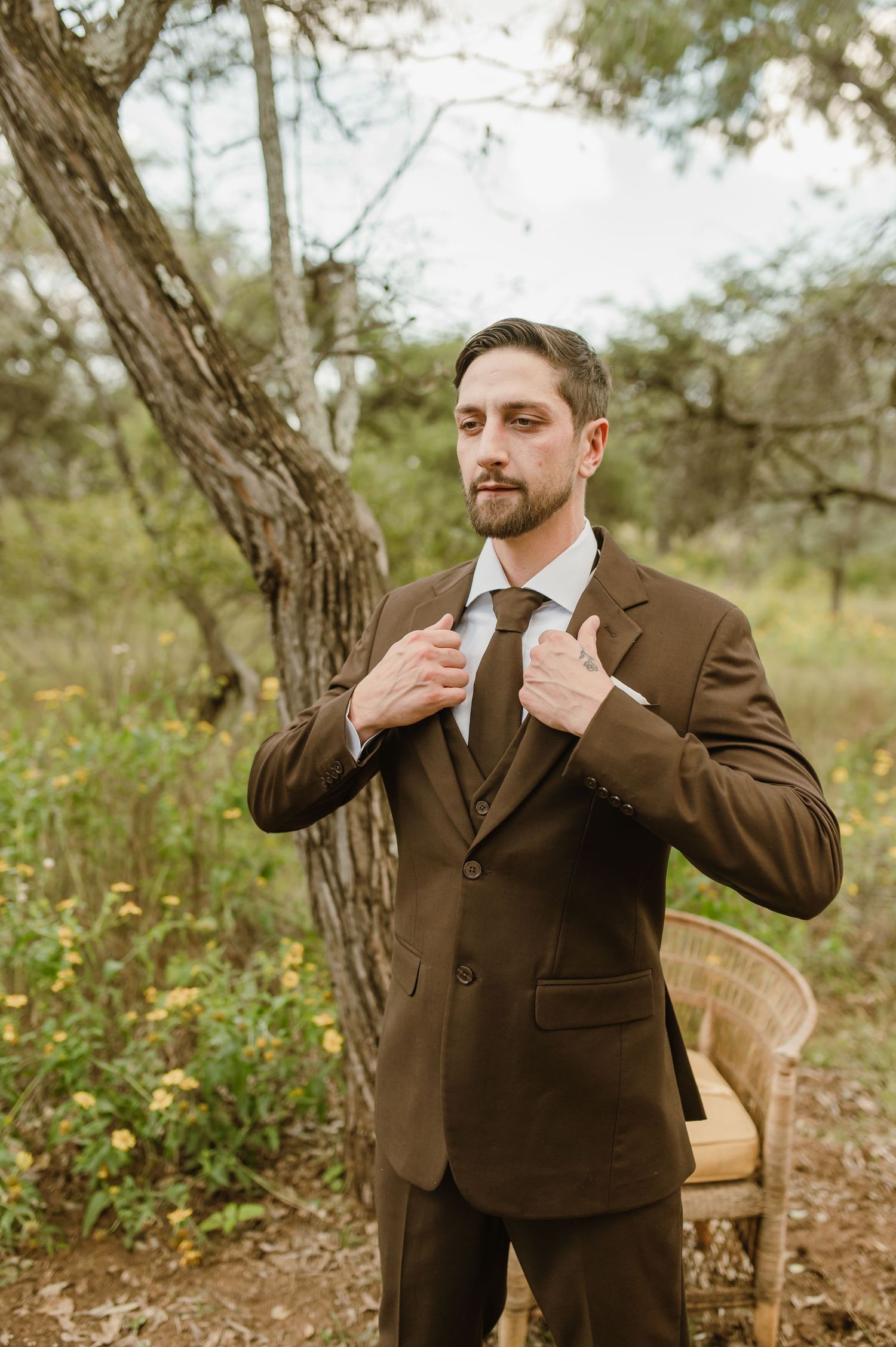Person adjusting brown suit and tie while standing in wooded outdoor setting.