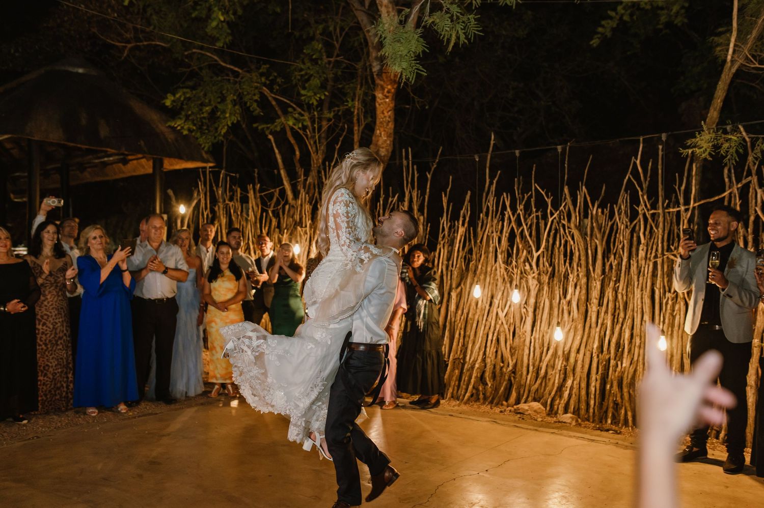 A couple dances energetically at their outdoor wedding reception with bamboo decor and string lights.