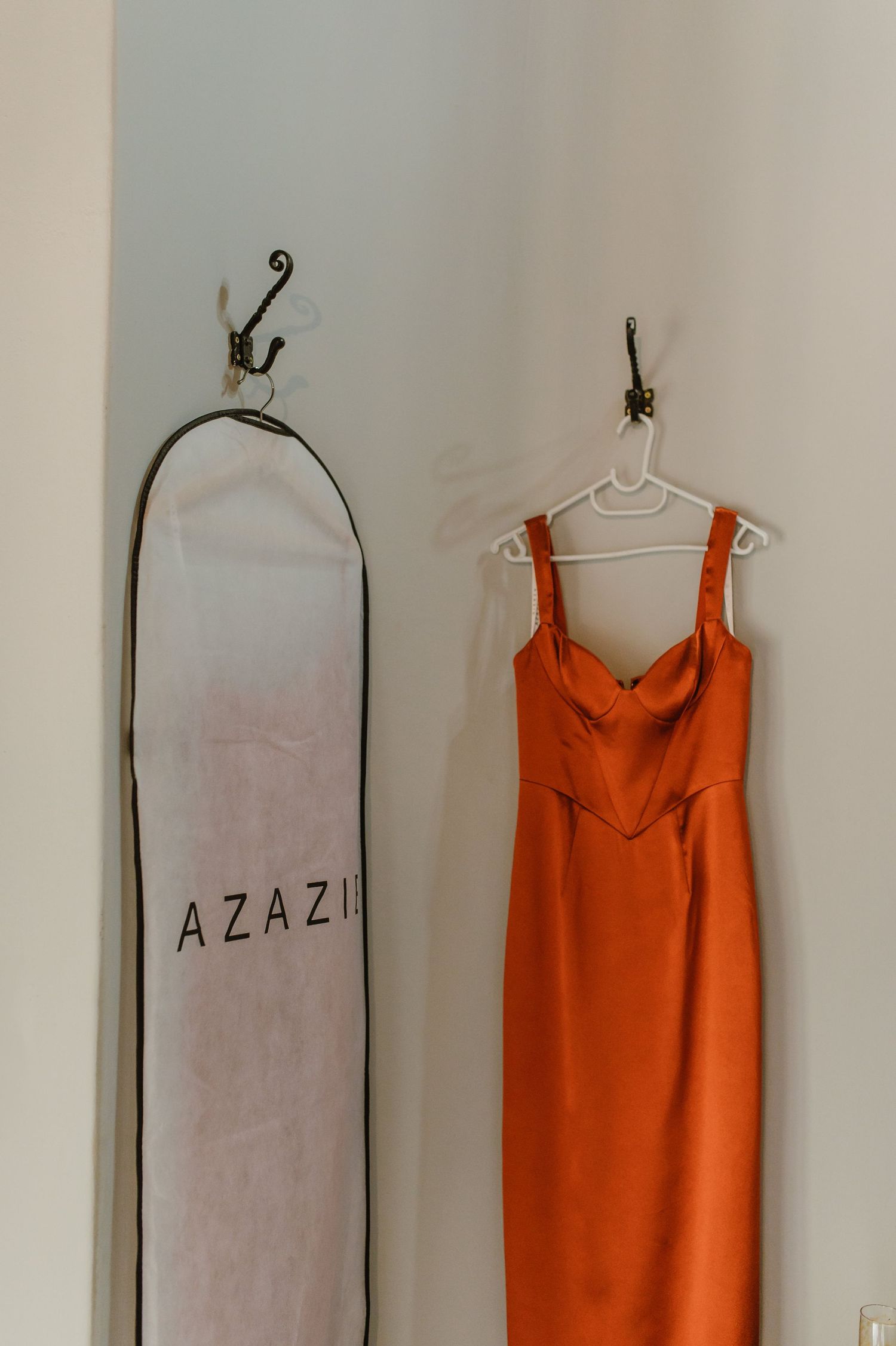 Orange bridesmaid dress hanging next to garment bags labeled 'AZAZIE' against white wall.