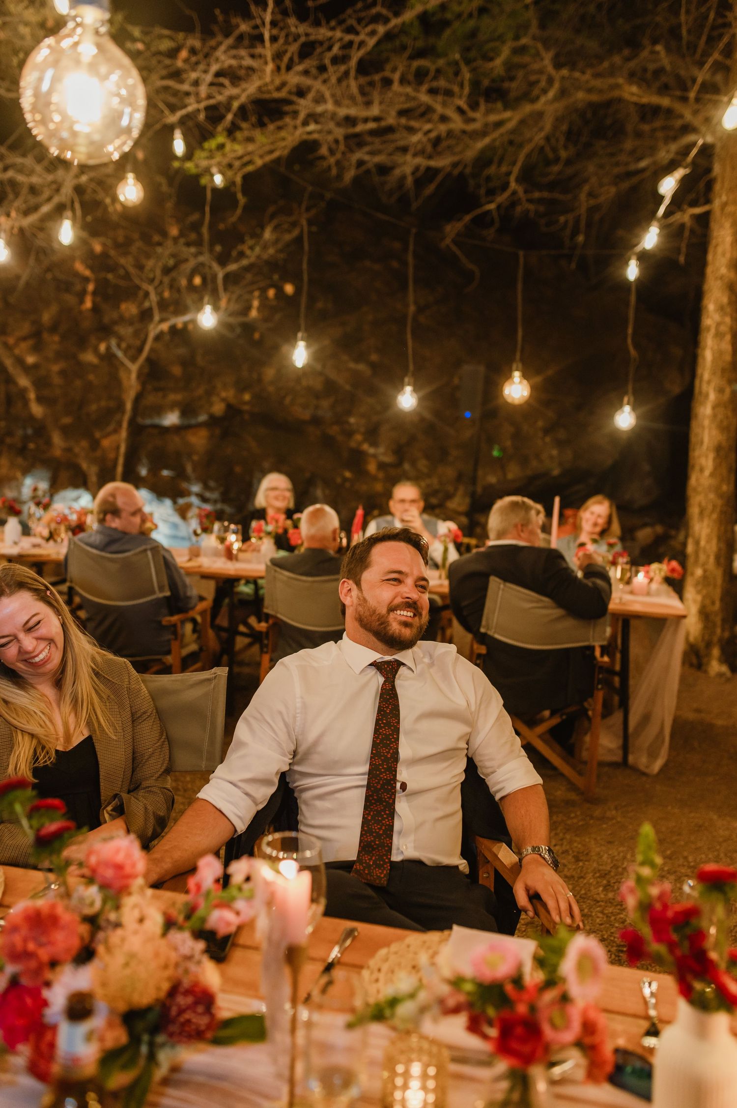 Wedding guests enjoying dinner under string lights with floral table decorations.