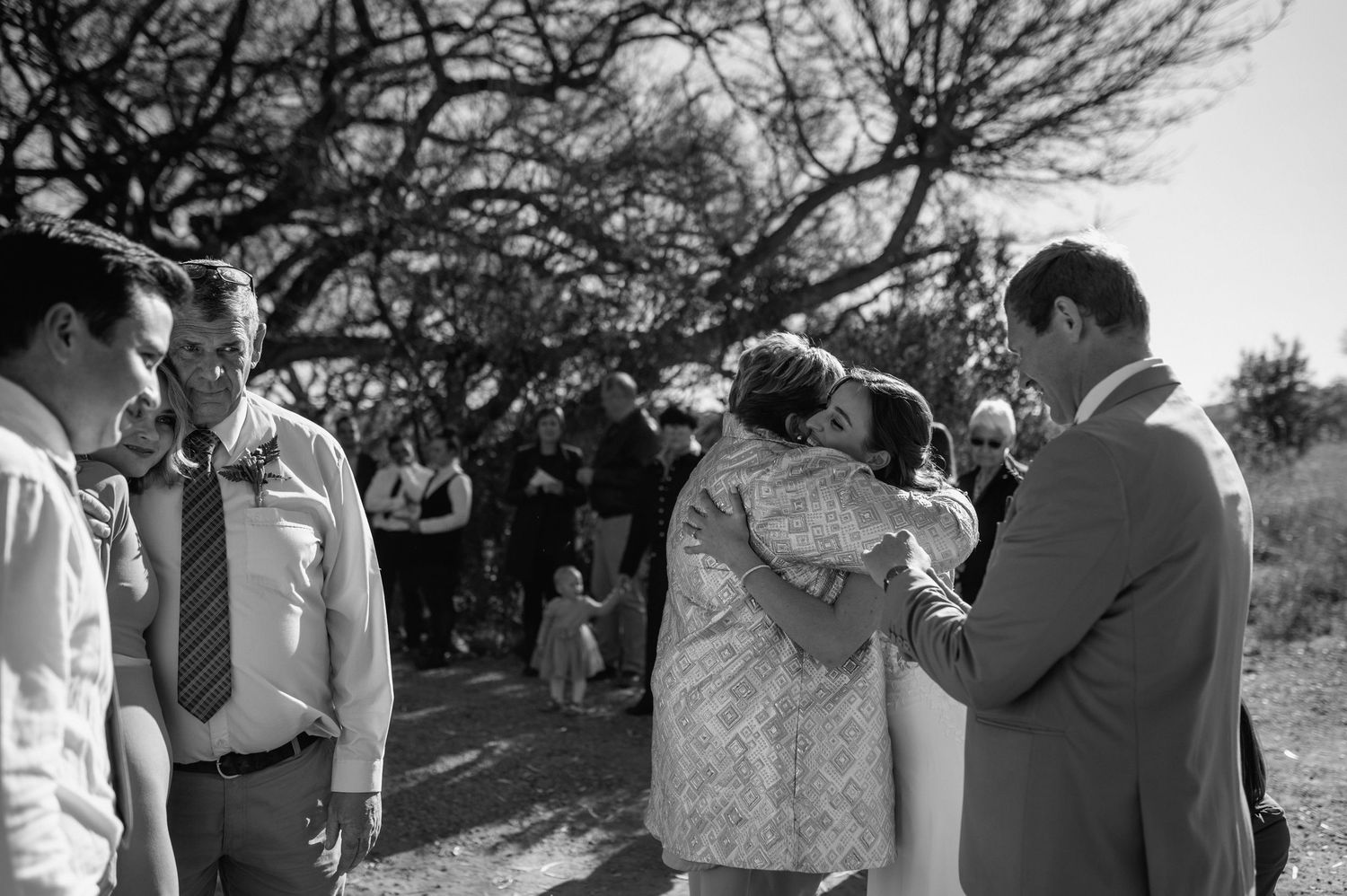 Wedding guests gather under trees during an outdoor ceremony in a natural setting.