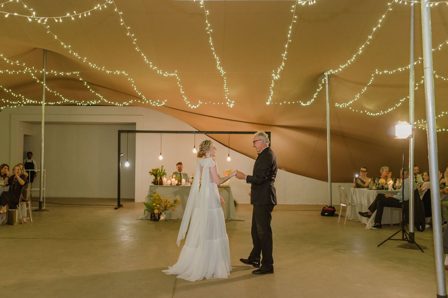 Wedding couple shares their first dance under twinkling string lights in an indoor venue.