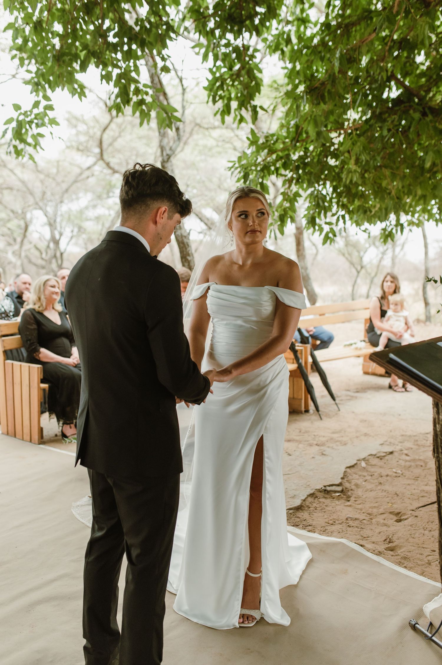 Intimate moment during outdoor wedding ceremony beneath green tree canopy.