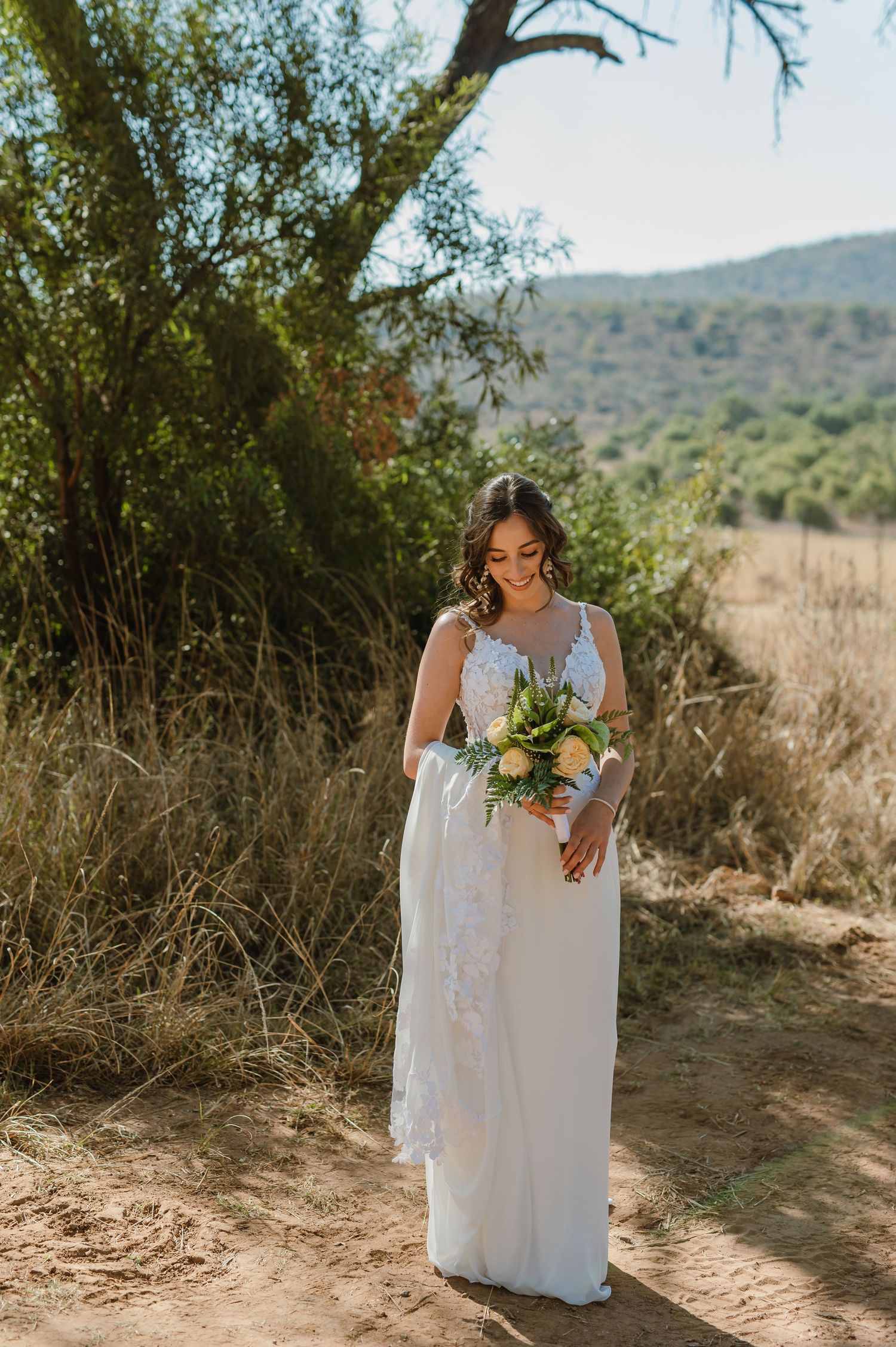 Bride in lace white gown holds bouquet while walking on desert trail.