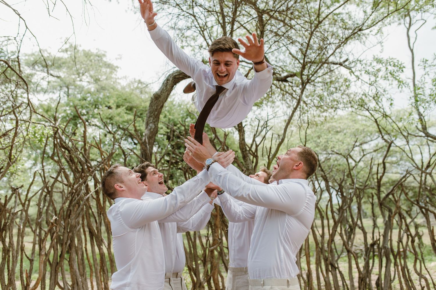 Wedding party members in light suits celebrate with energetic poses in a wooded area.