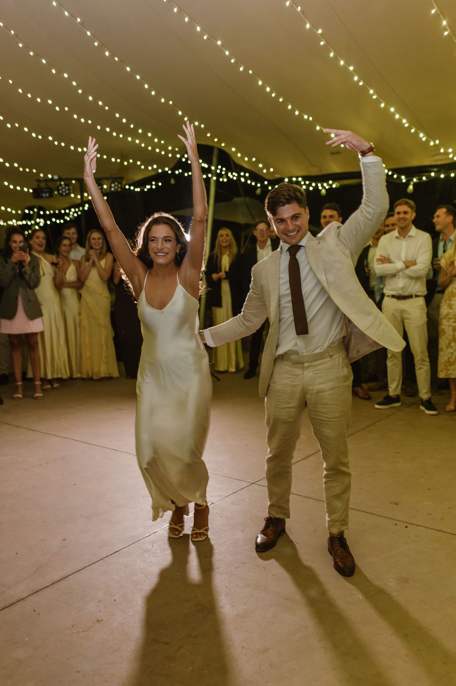 Dancers hold hands and lean back in joyful celebration under tent lighting at a wedding reception.