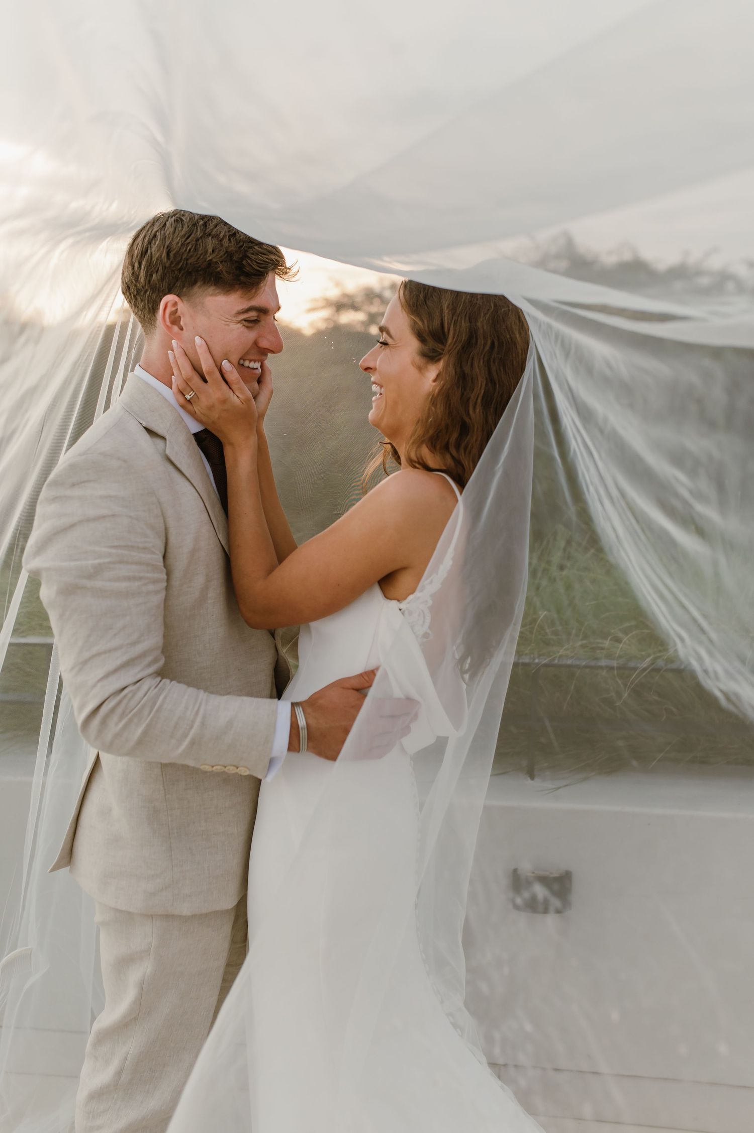 Wedding couple embraces under flowing white veil with natural scenery in background.