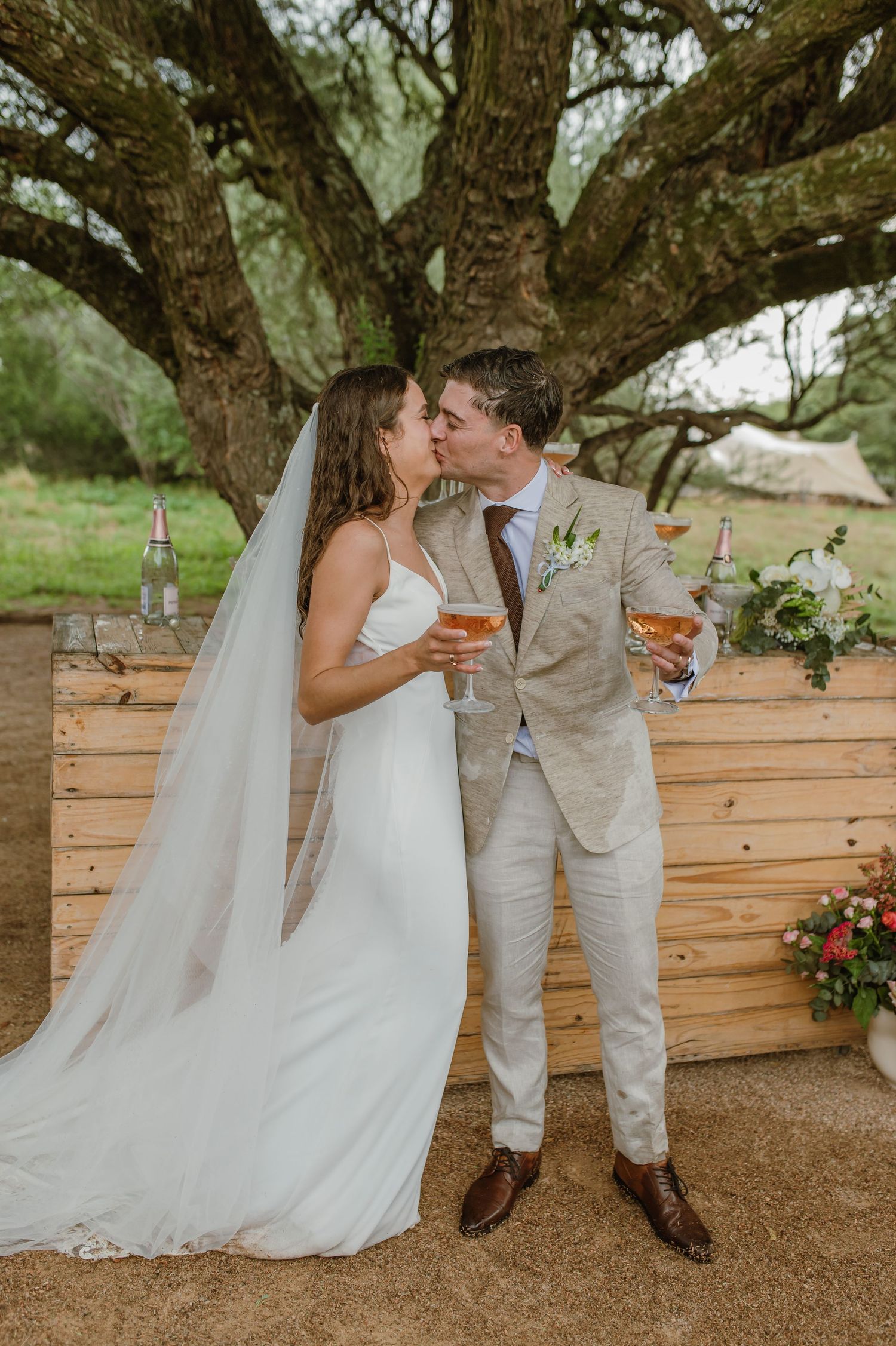 Wedding couple enjoying drinks together against a rustic wooden backdrop under a tree.