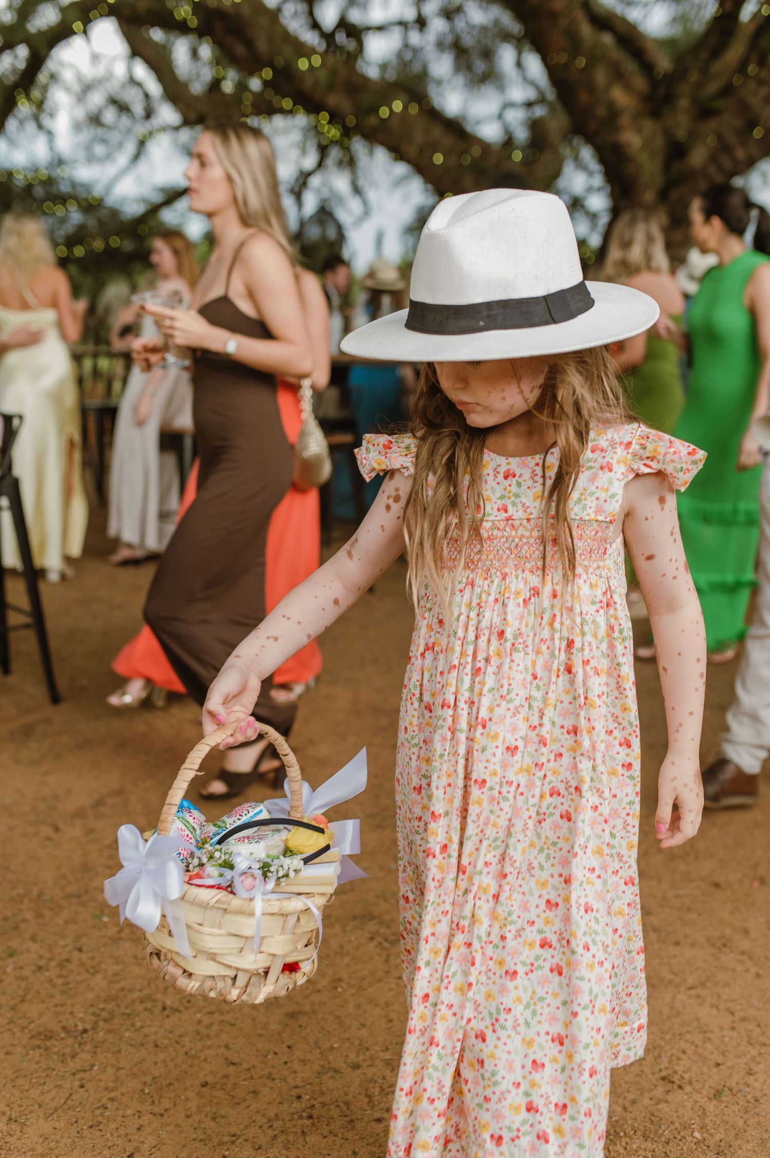 Guest in a floral dress and sun hat carrying a basket at an outdoor event.