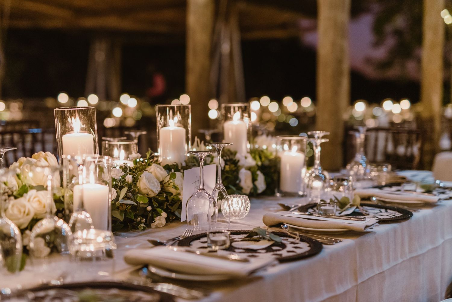 Long reception tables decorated with candlelight and floral arrangements under rustic wooden beams.