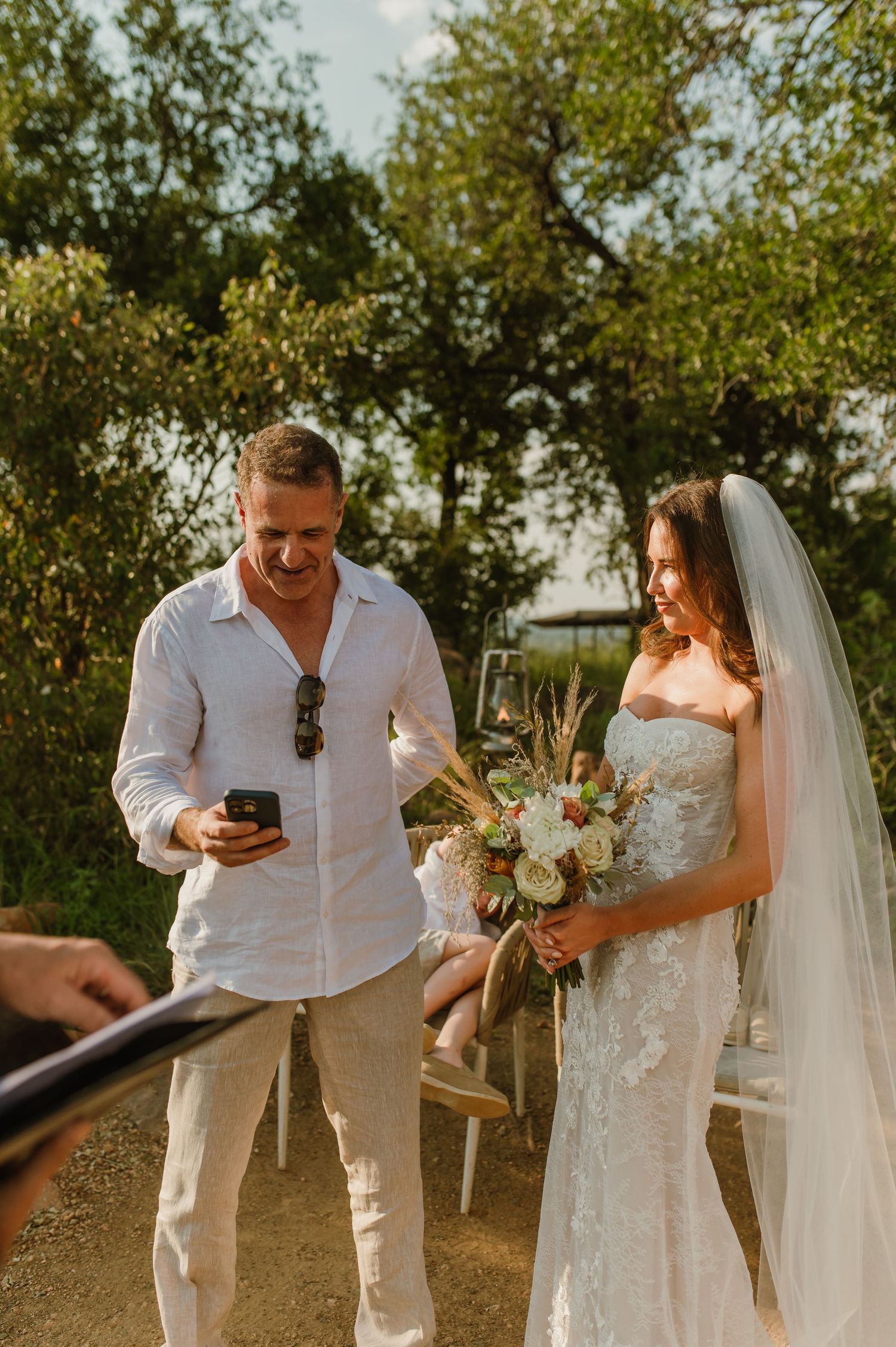 Bride in strapless lace gown holds bouquet beside smiling man in white at outdoor wedding ceremony.