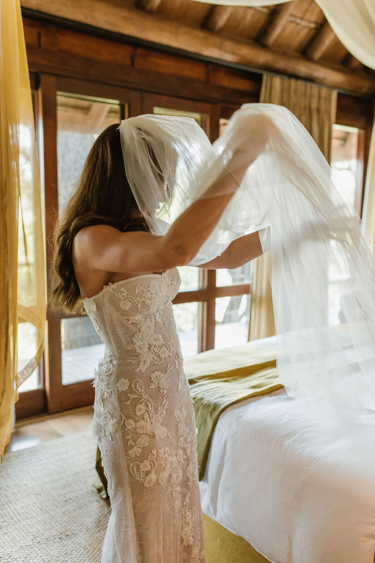 Bride in strapless lace wedding gown placing a veil in a rustic wooden lodge bedroom.