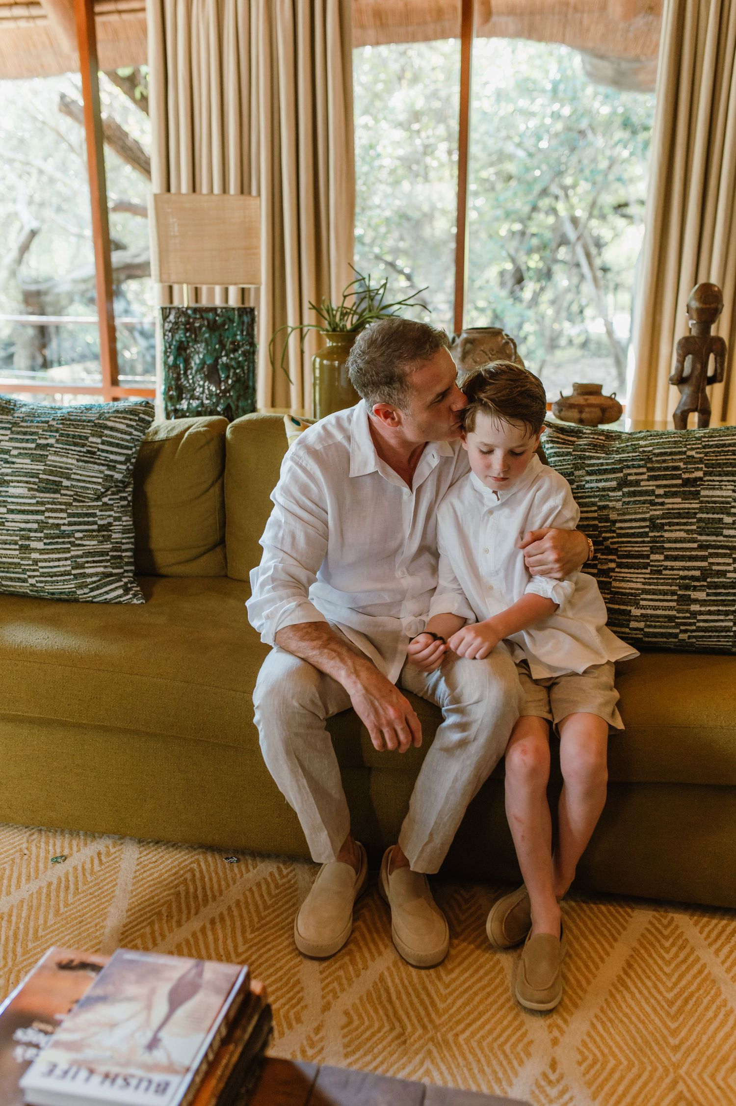 Father and son sharing tender embrace on olive green sofa in cozy safari lodge living room.