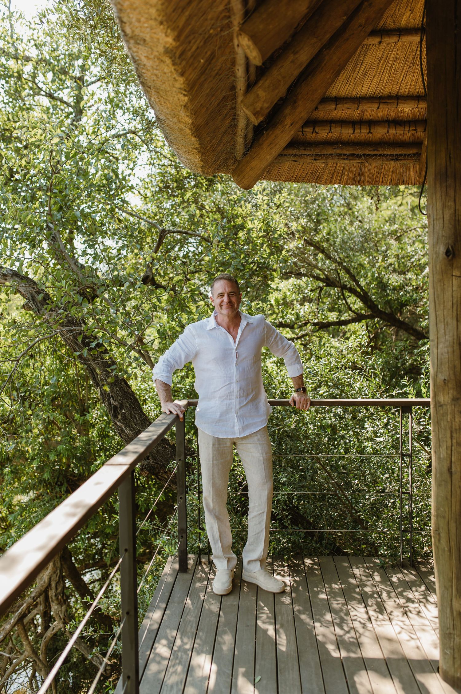 Smiling man in white shirt and cream pants posing on wooden treehouse balcony surrounded by lush green trees.