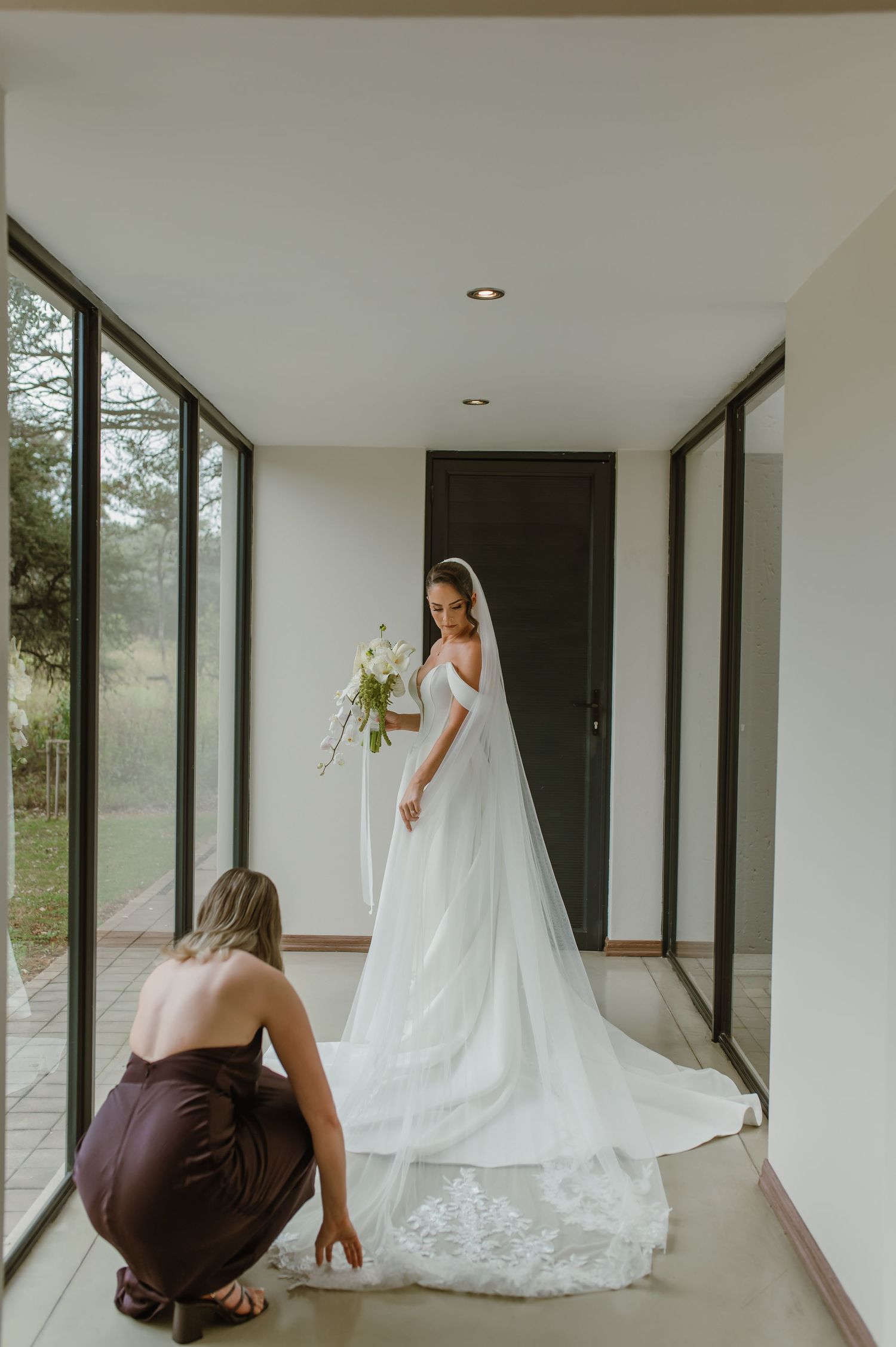 Wedding dress fitting scene in modern room with floor-length gown and flowing veil being adjusted.