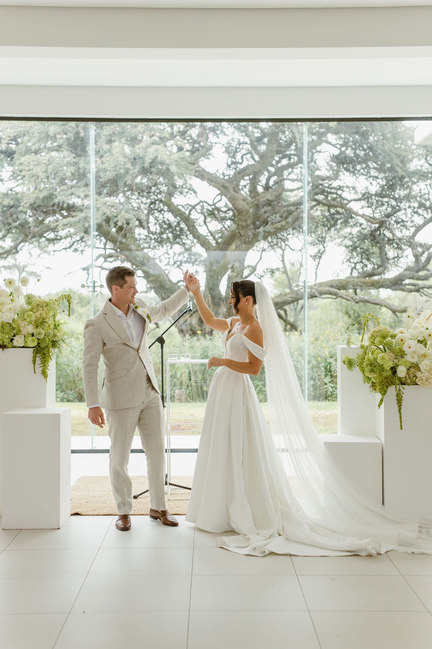 A bride and groom celebrate during their ceremony in a bright, modern venue with large windows and floral decor.