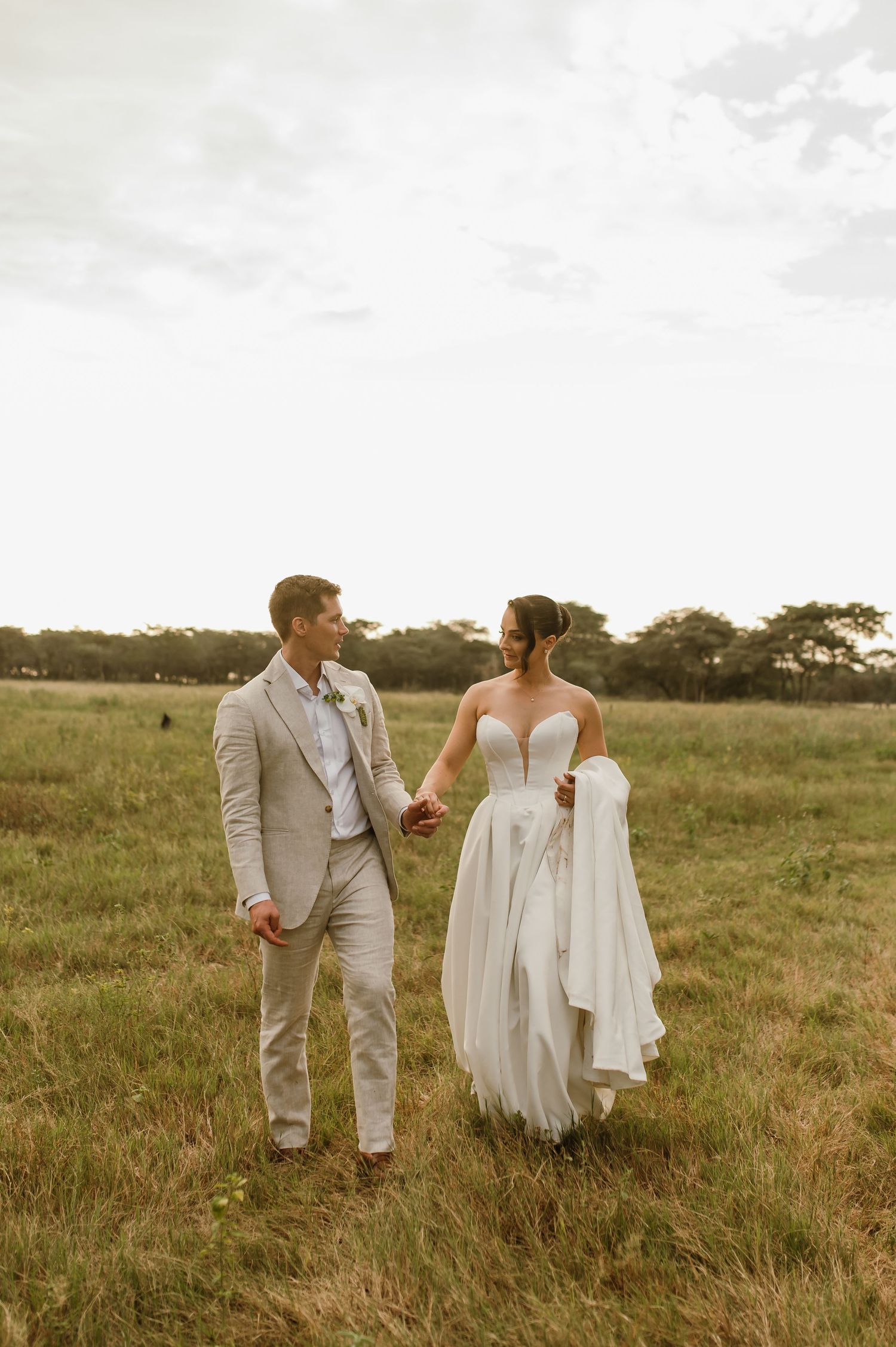 A couple in wedding attire walking together through a golden field at sunset.
