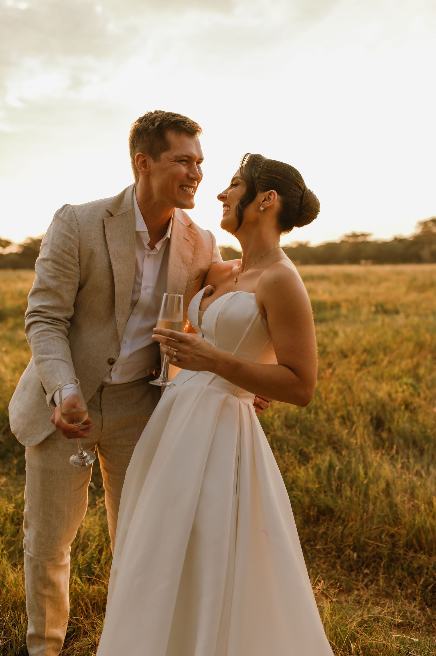 A wedding couple shares champagne and laughter in a sun-drenched field at sunset.