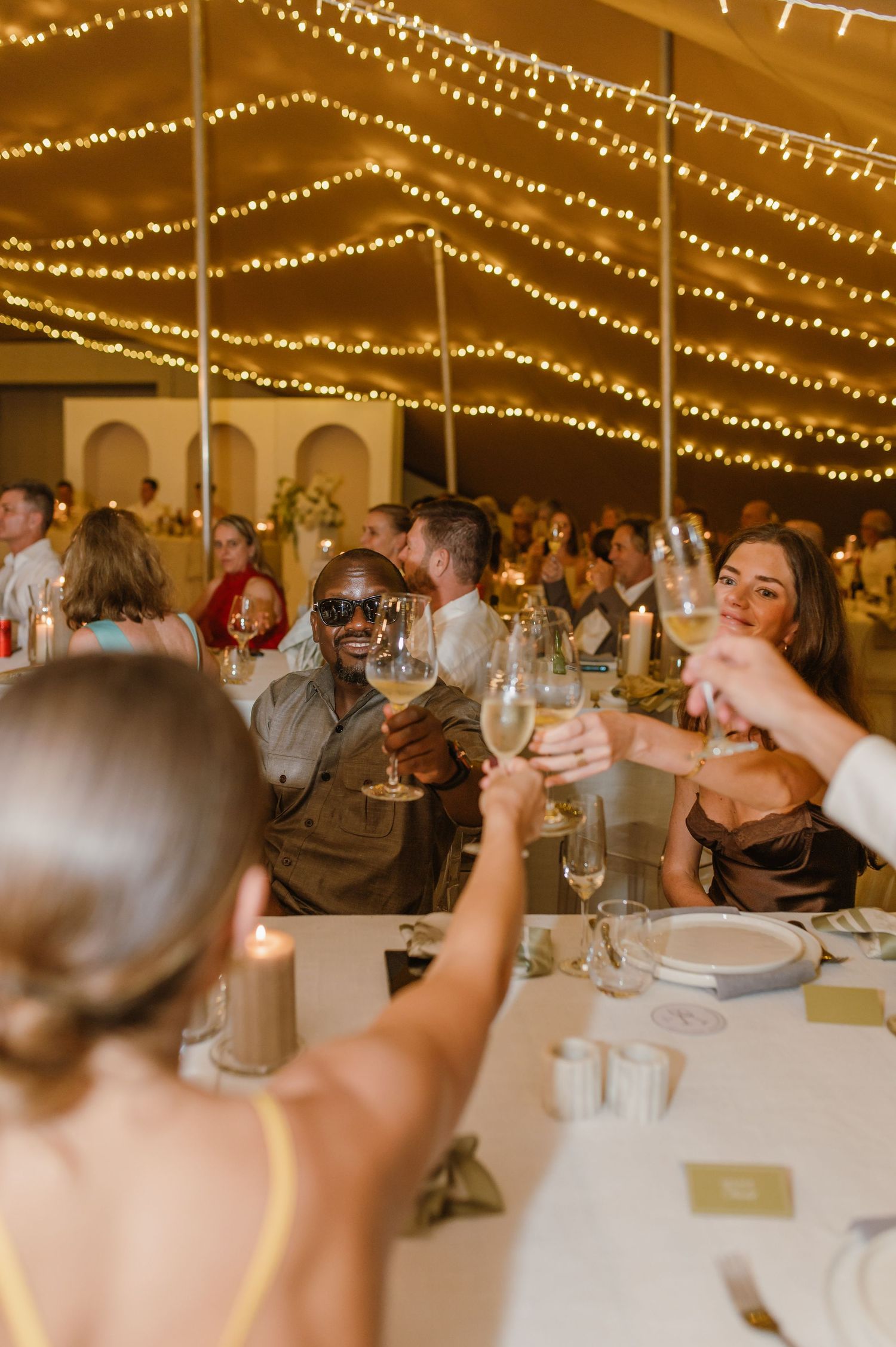 Wedding reception guests toasting under warm string lights in an elegant tent setting.