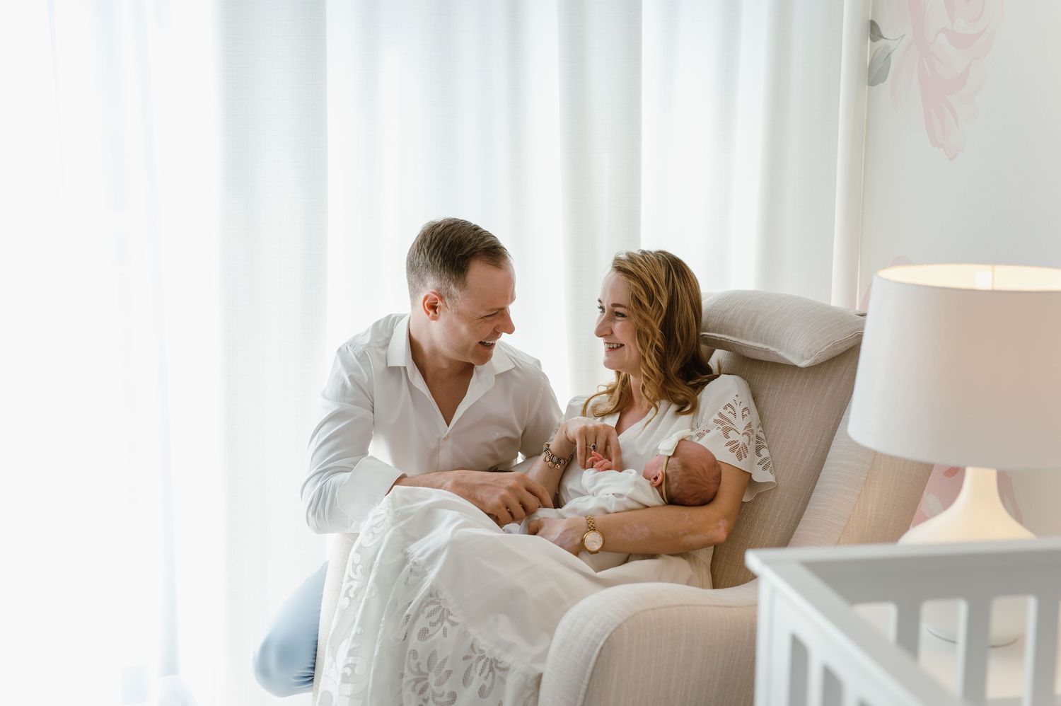 A couple in white clothing relaxes and laughs together on a cozy bed with white linens.