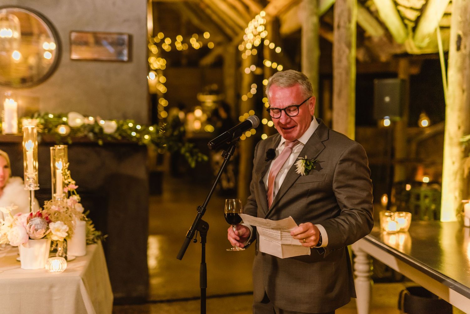 Wedding guests seated at long tables decorated with candles and floral arrangements in a rustic venue.