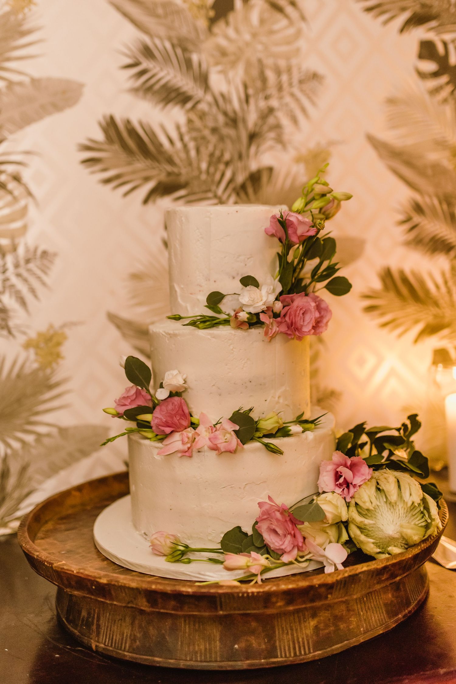 White tiered wedding cake decorated with pink roses and greenery on a wooden stand.