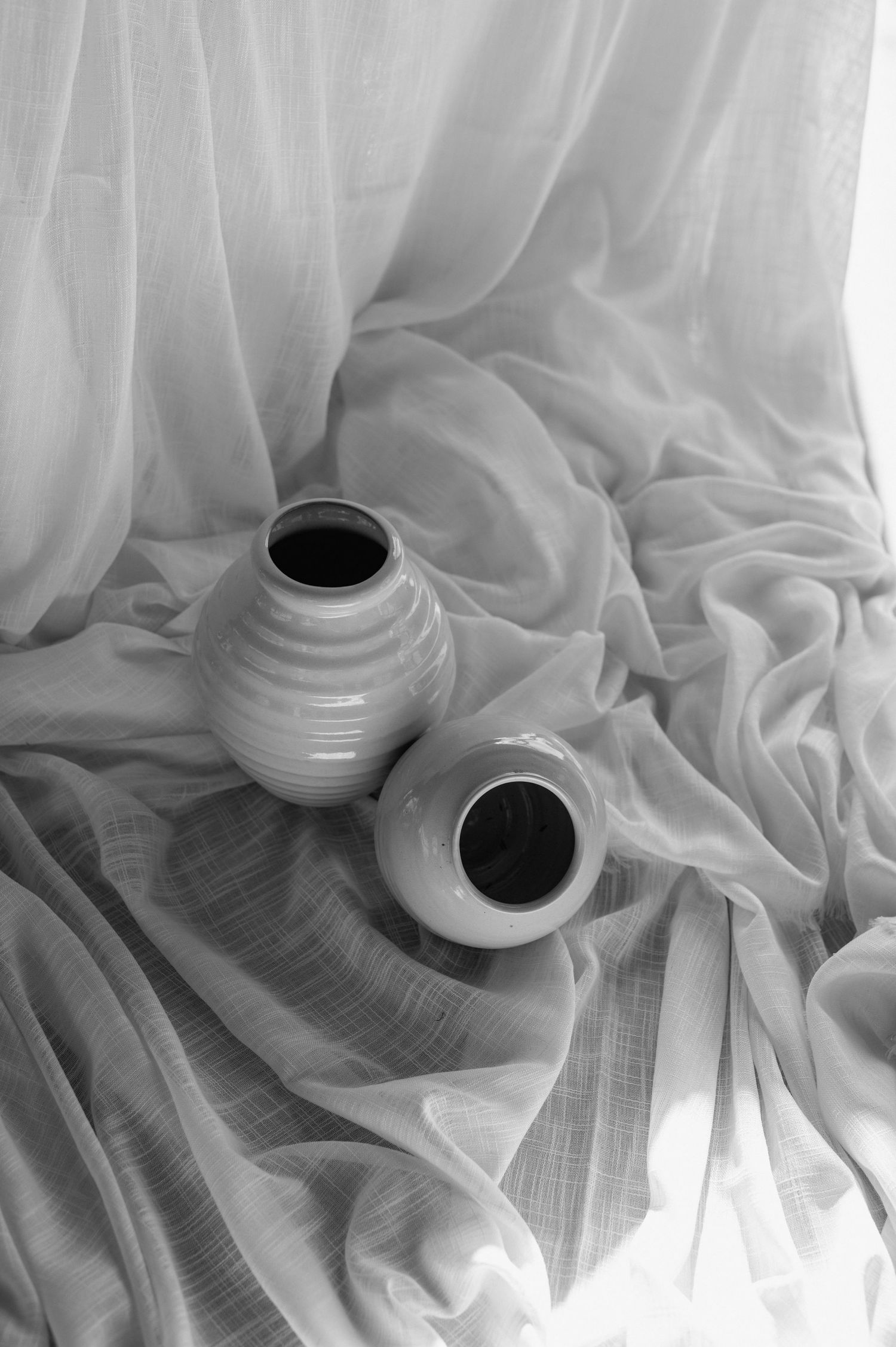 Black and white still life of two ceramic vases against flowing white fabric.