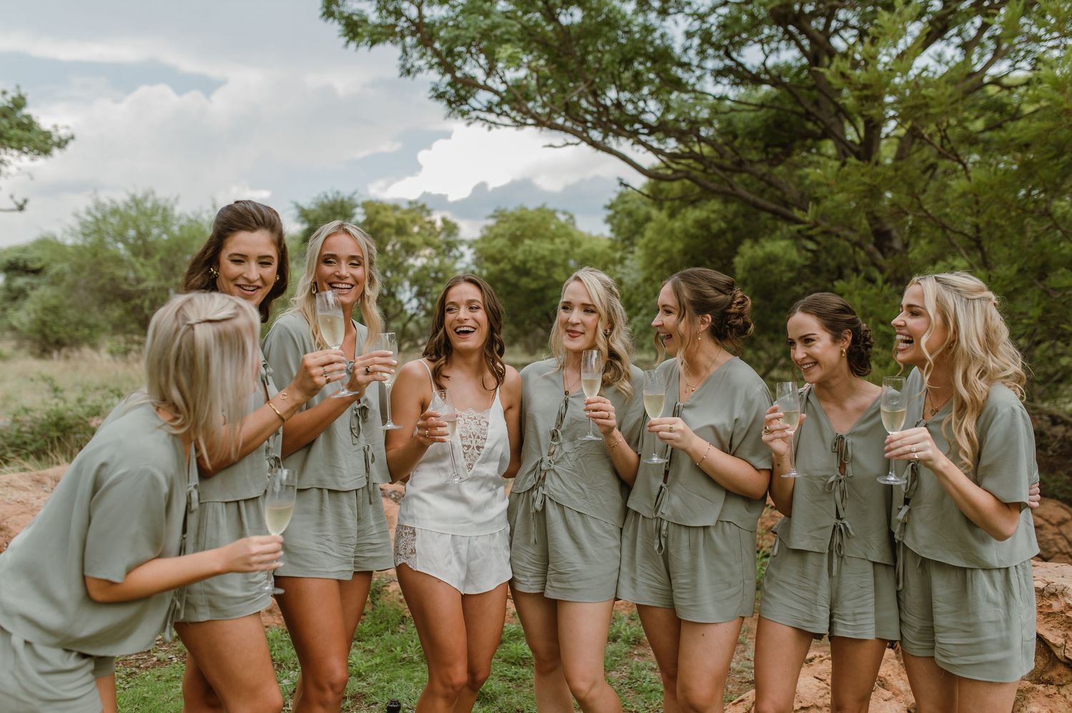 Group wearing matching sage outfits toasts with champagne glasses in an outdoor garden setting.