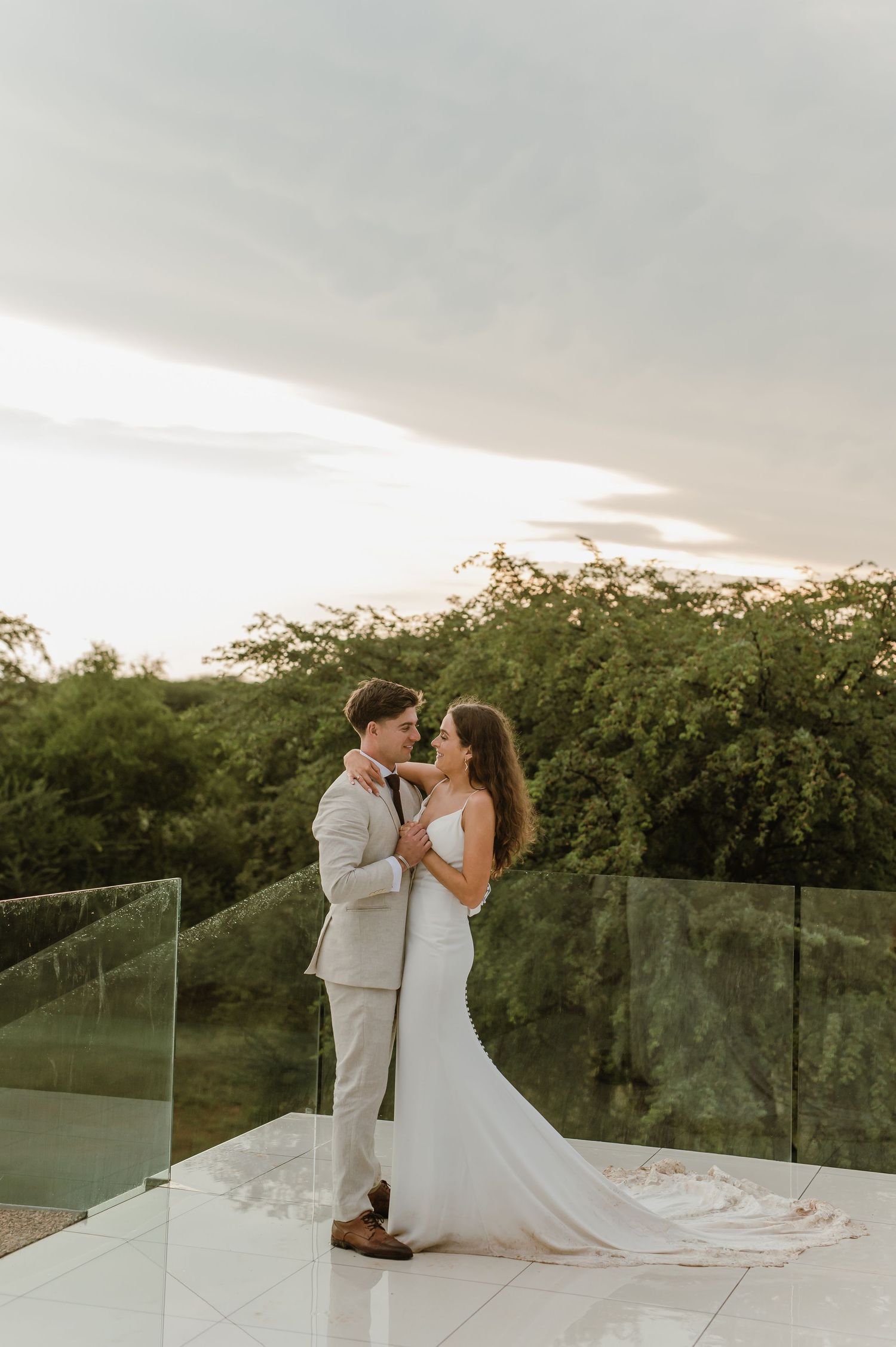 A romantic wedding couple embraces on a scenic outdoor terrace at sunset.