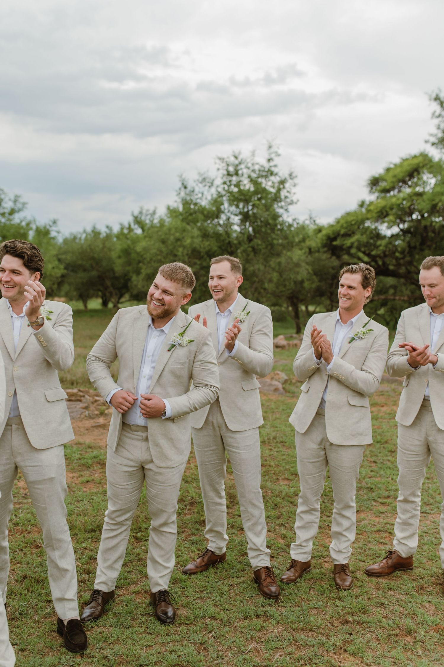 Groomsmen in beige suits posing playfully in a line outdoors.