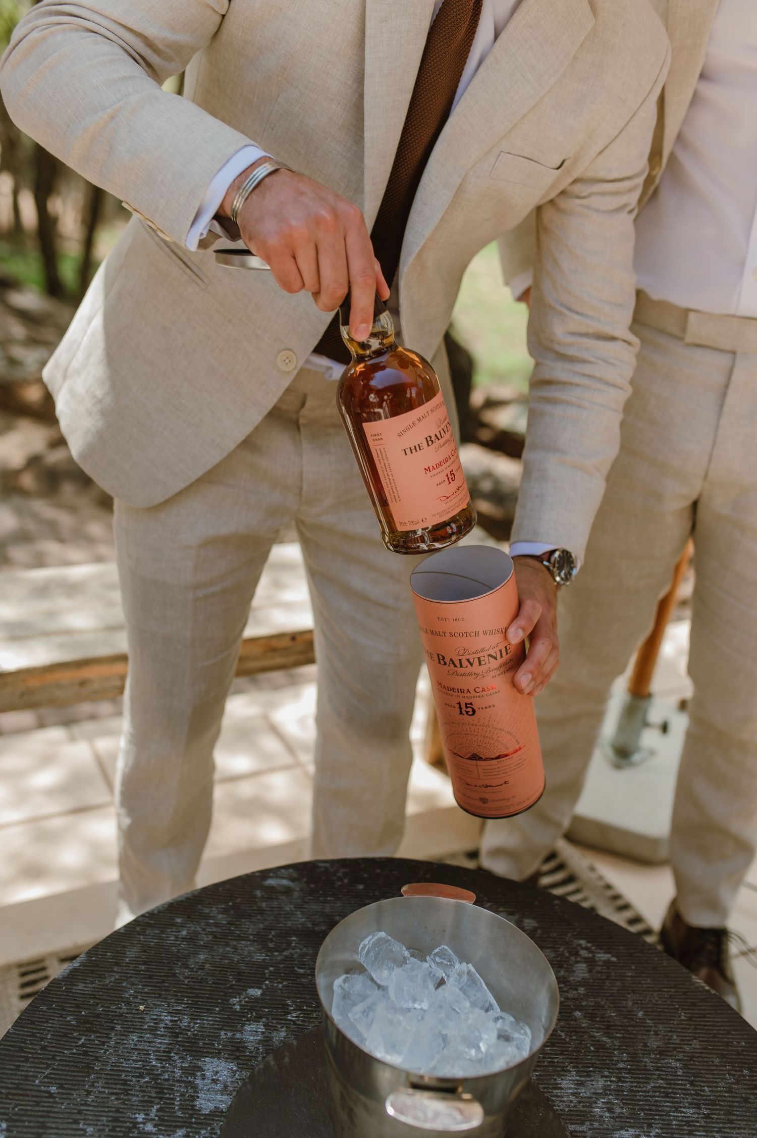 Groomsmen in beige suits pour drinks over ice at an outdoor celebration.