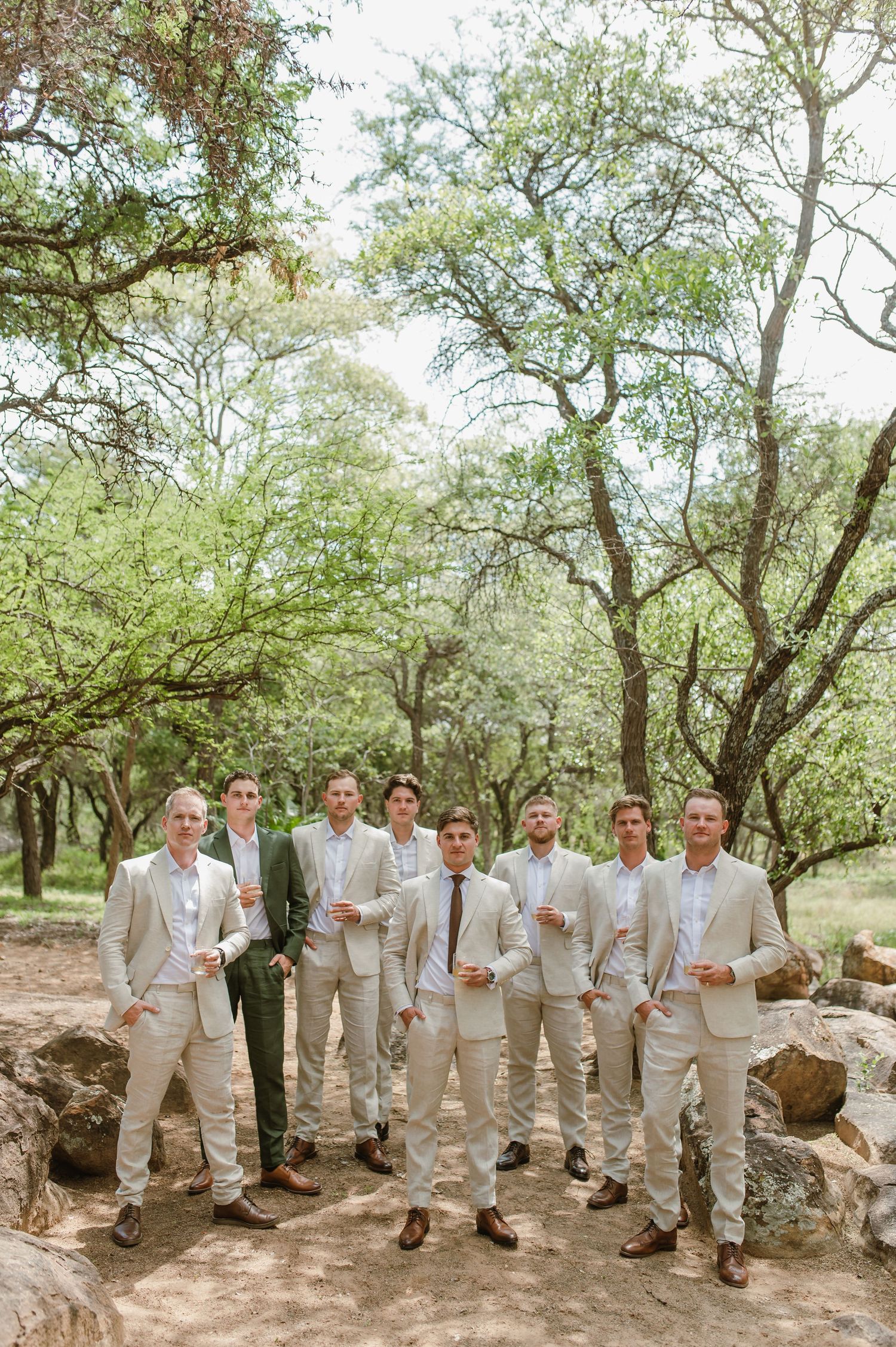 Large wedding party in coordinated beige suits poses together among trees.