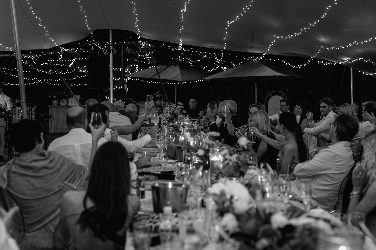 Black and white photo of a wedding reception under string lights with guests seated at decorated tables.
