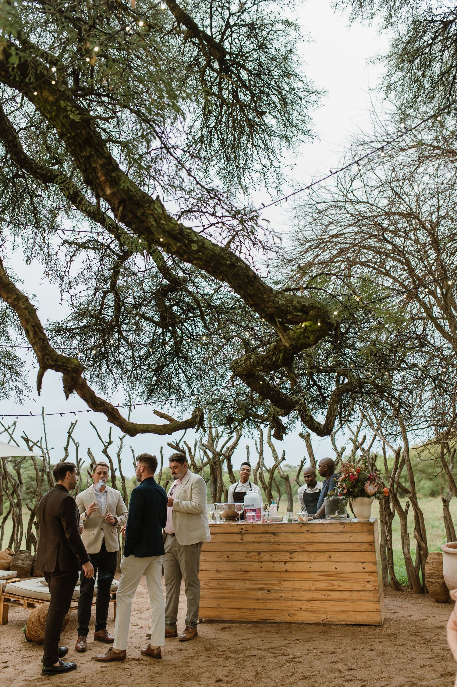 Wedding guests mingling around a wooden bar setup under large trees.