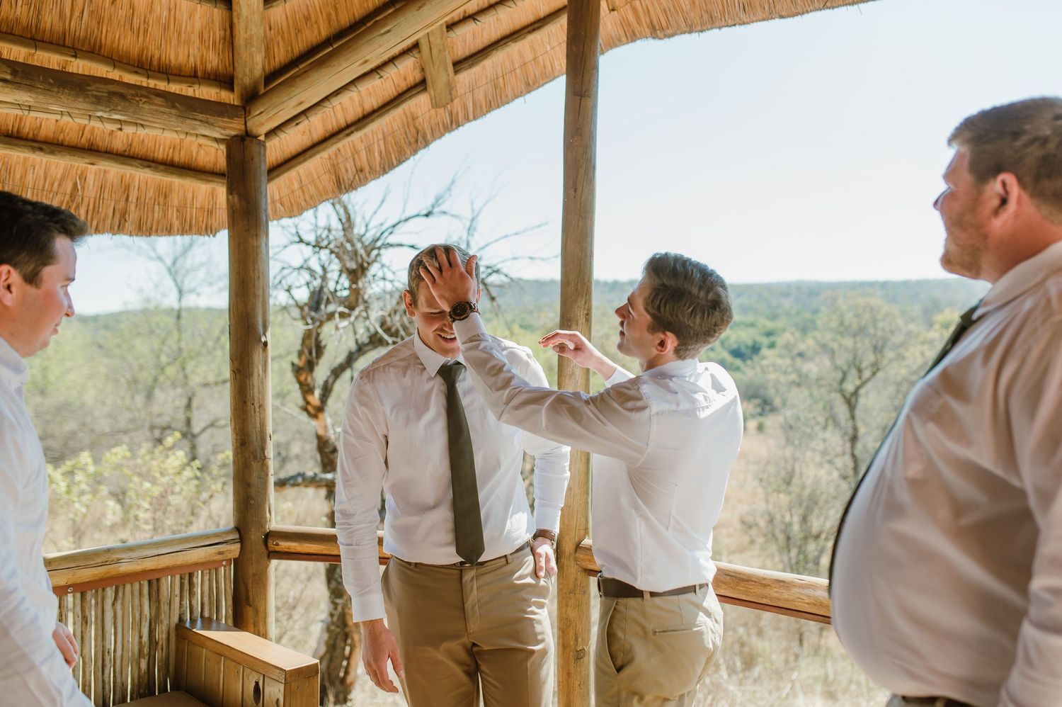 Groomsmen gather on a rustic covered deck overlooking scenic African wilderness landscape.