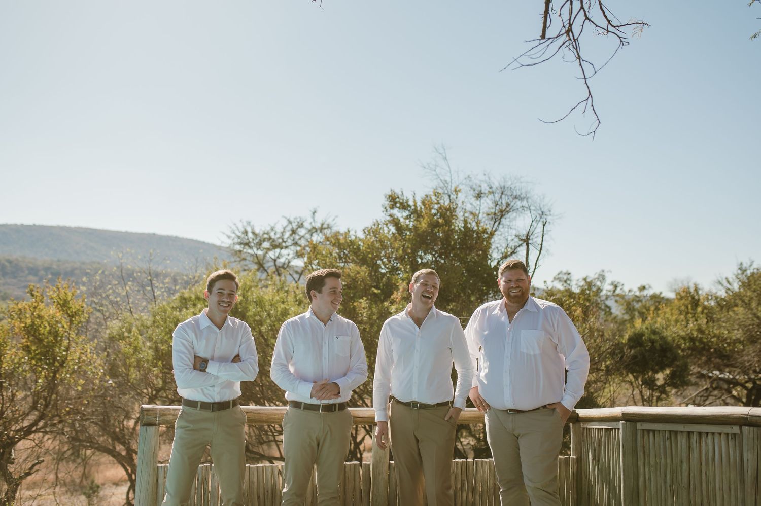 Groomsmen in matching outfits standing against railing with scenic mountain backdrop.