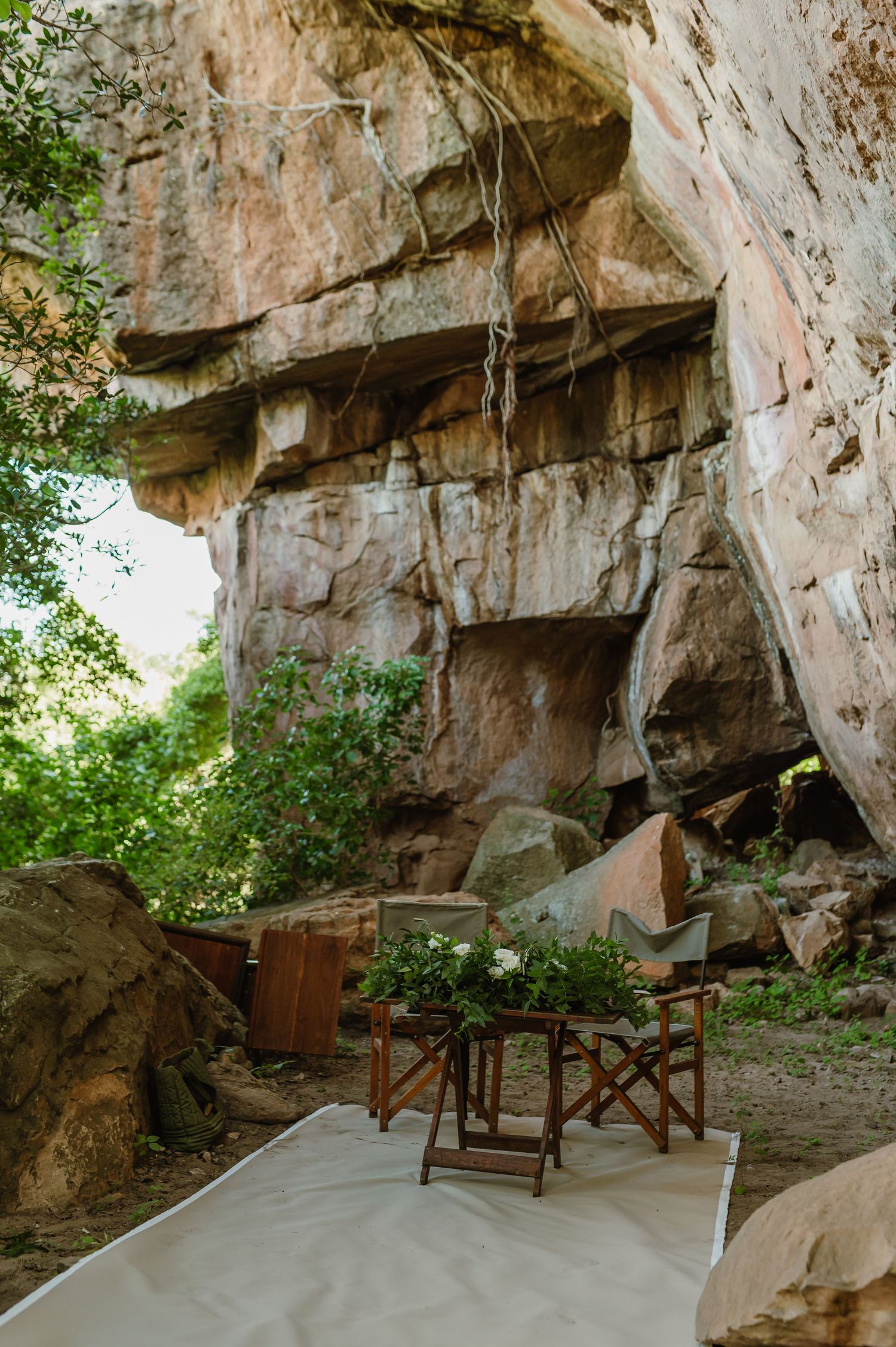 Rustic picnic tables arranged under dramatic rock overhang for outdoor wedding reception.