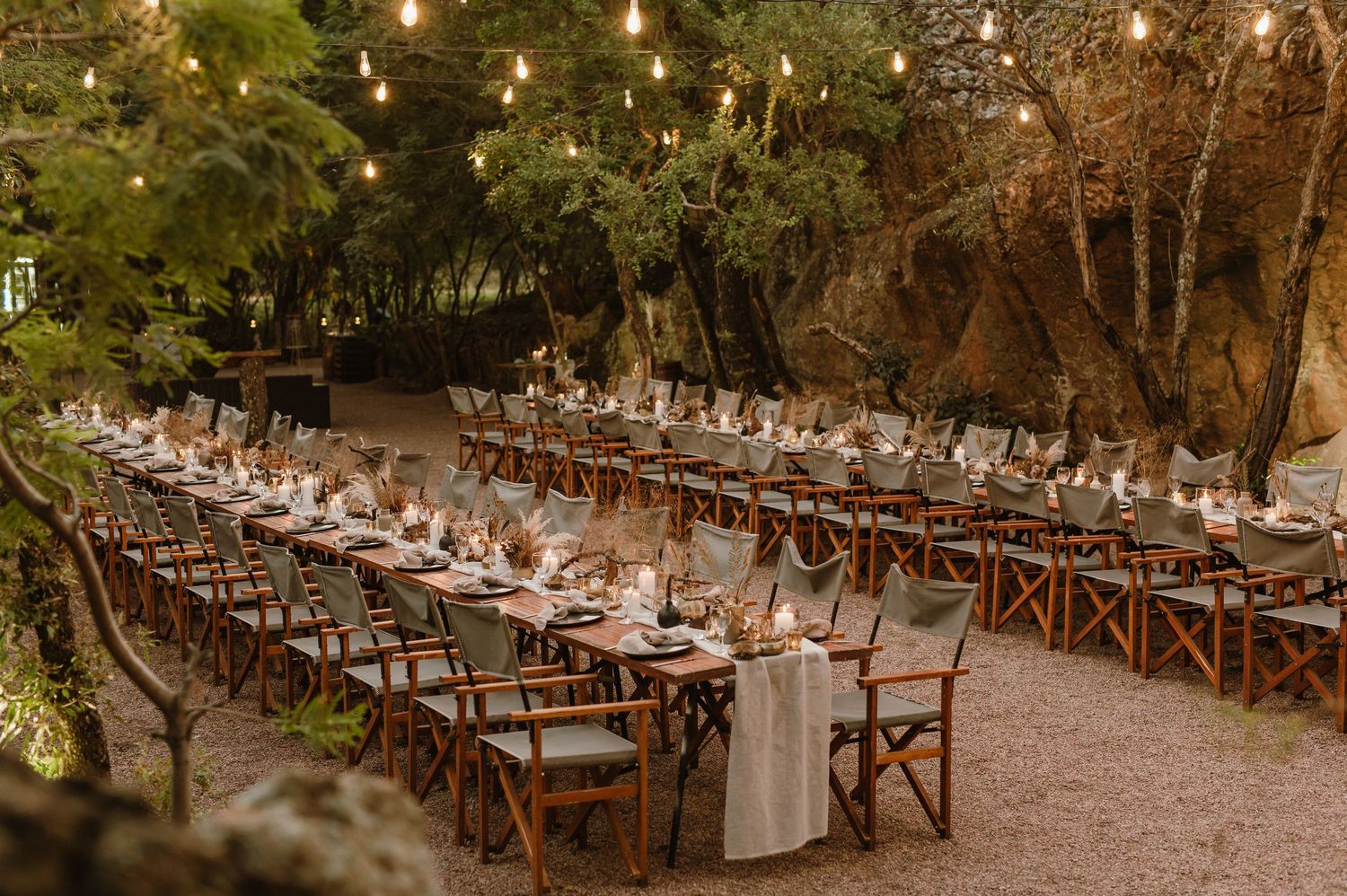 Long rustic wooden tables set for an elegant outdoor dinner party under string lights and trees.