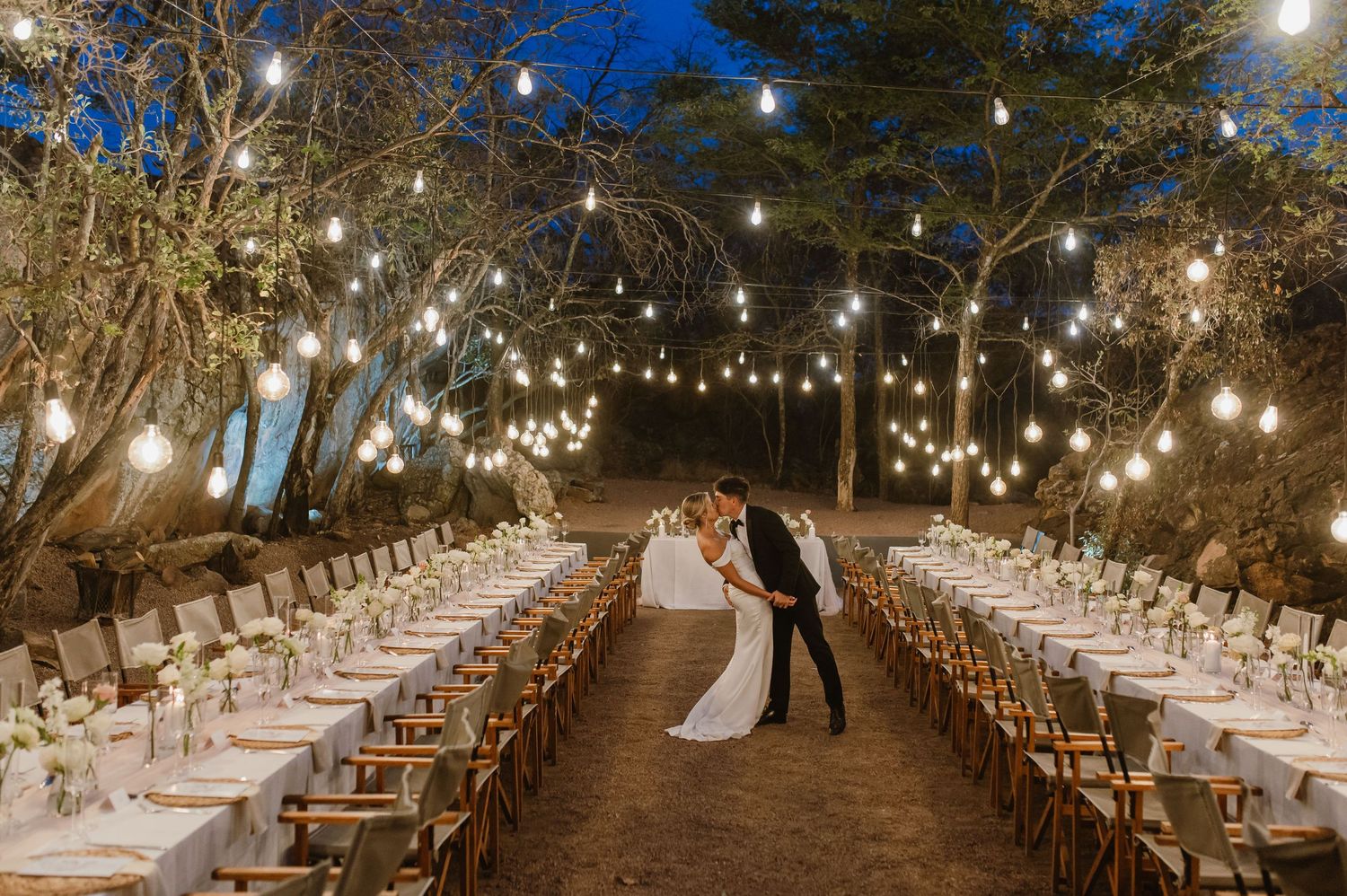 Enchanting outdoor wedding reception space with rows of tables under canopy of glowing string lights.